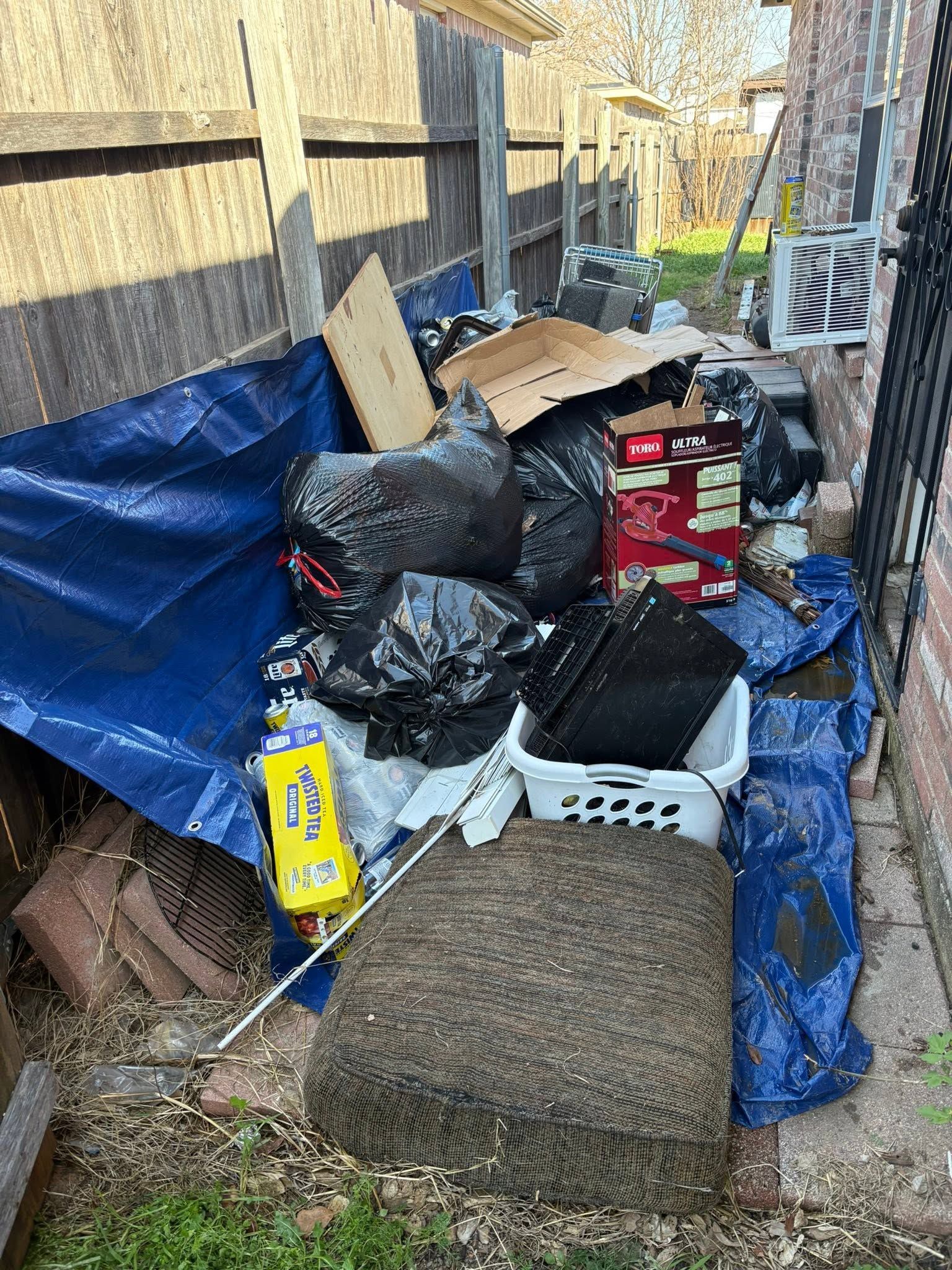 Cluttered outdoor storage area with blue tarp, stacked boxes, bags, and debris beside a fence and building.