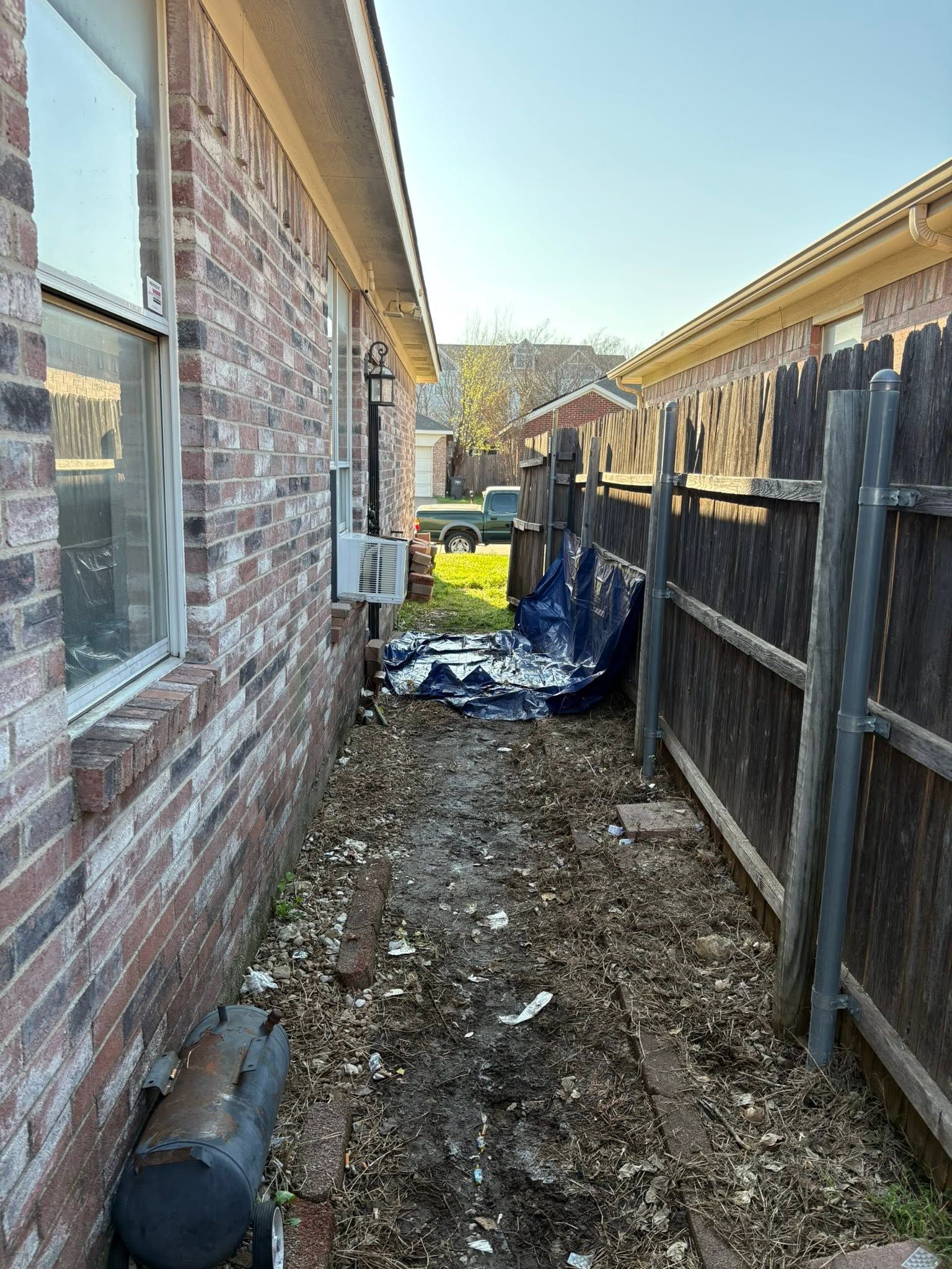 Narrow side yard between brick house and wooden fence, with debris and a blue tarp on the ground