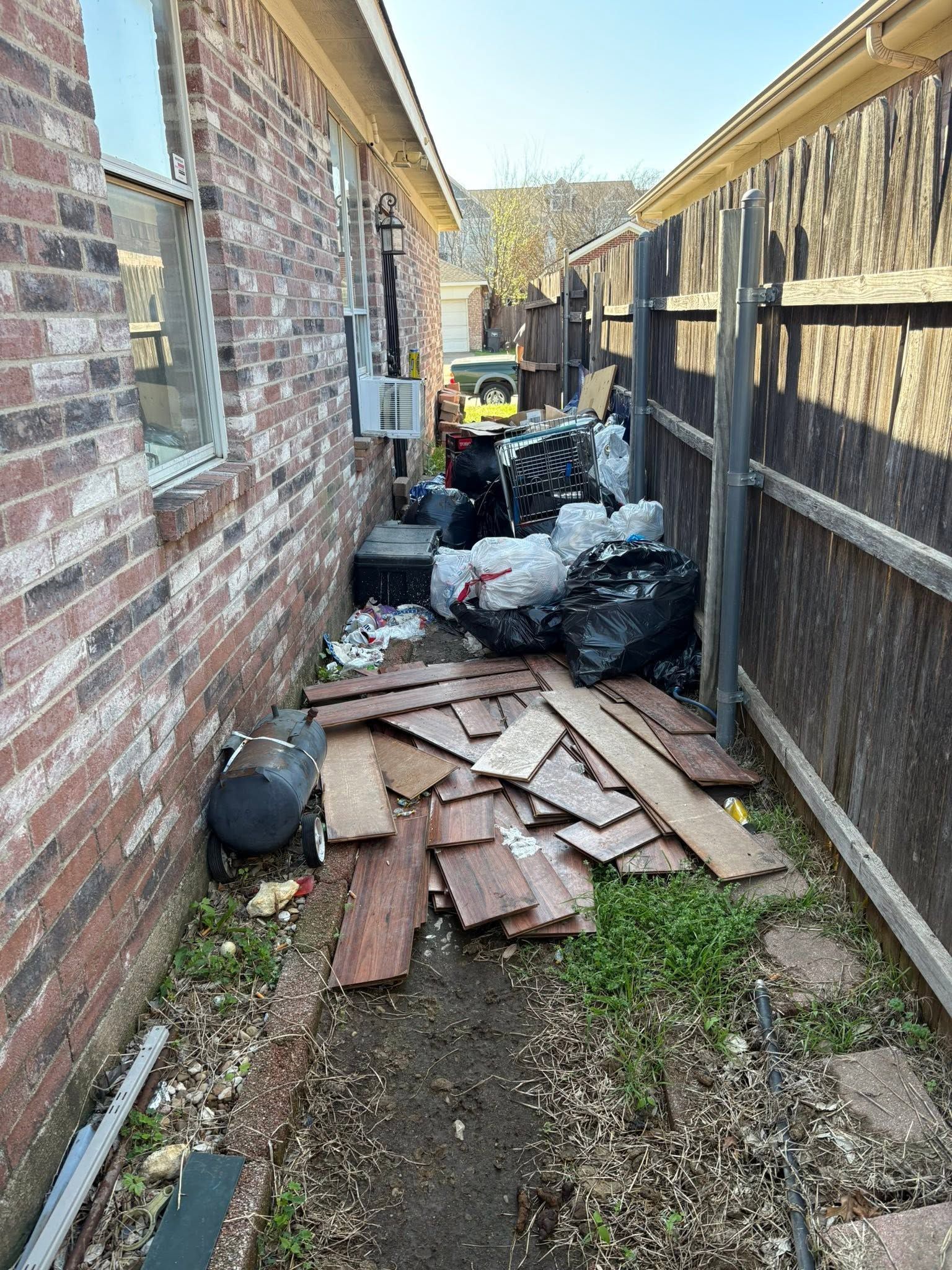 Narrow alley beside brick and wooden fences with scattered trash bags, boards, and debris on the ground.