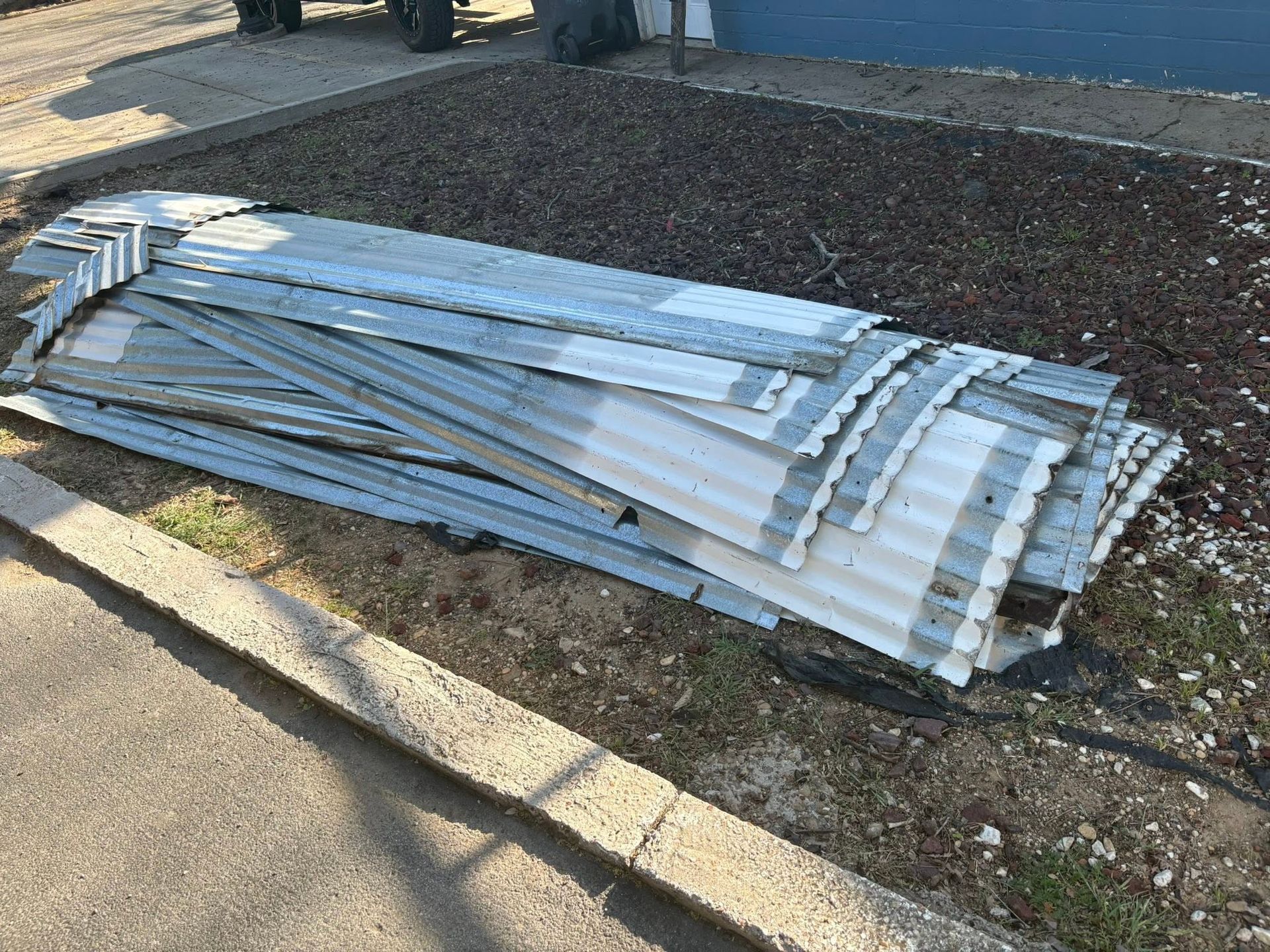 Stack of bent white metal panels lying on a landscaped curb beside a sidewalk.