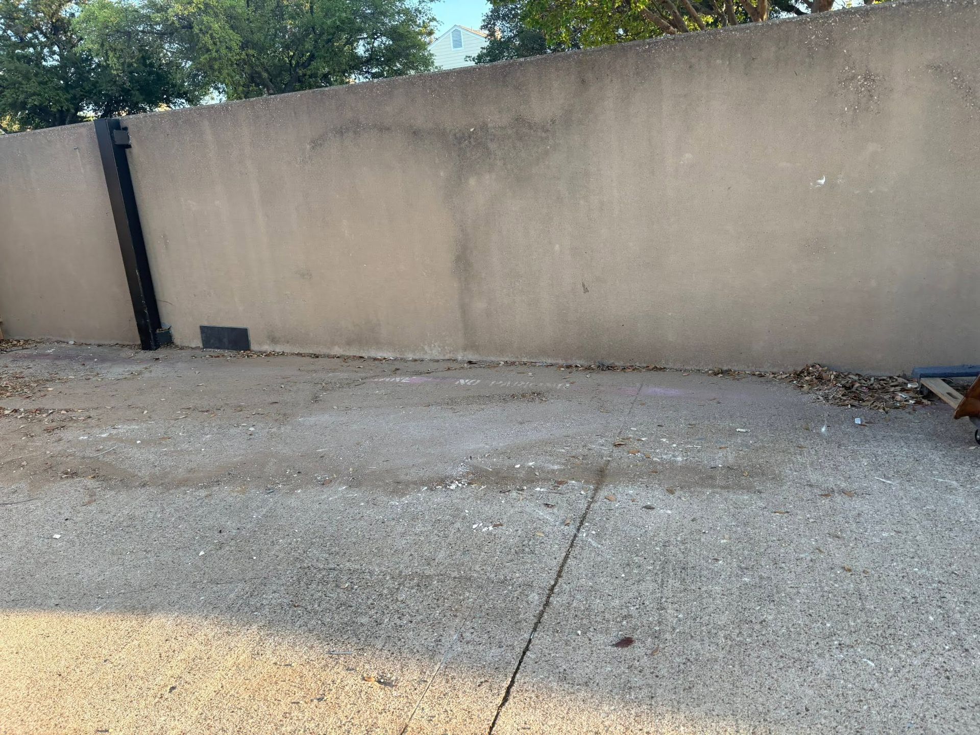 Concrete driveway beside a tan stucco wall with a black gate, lit by sunlight.