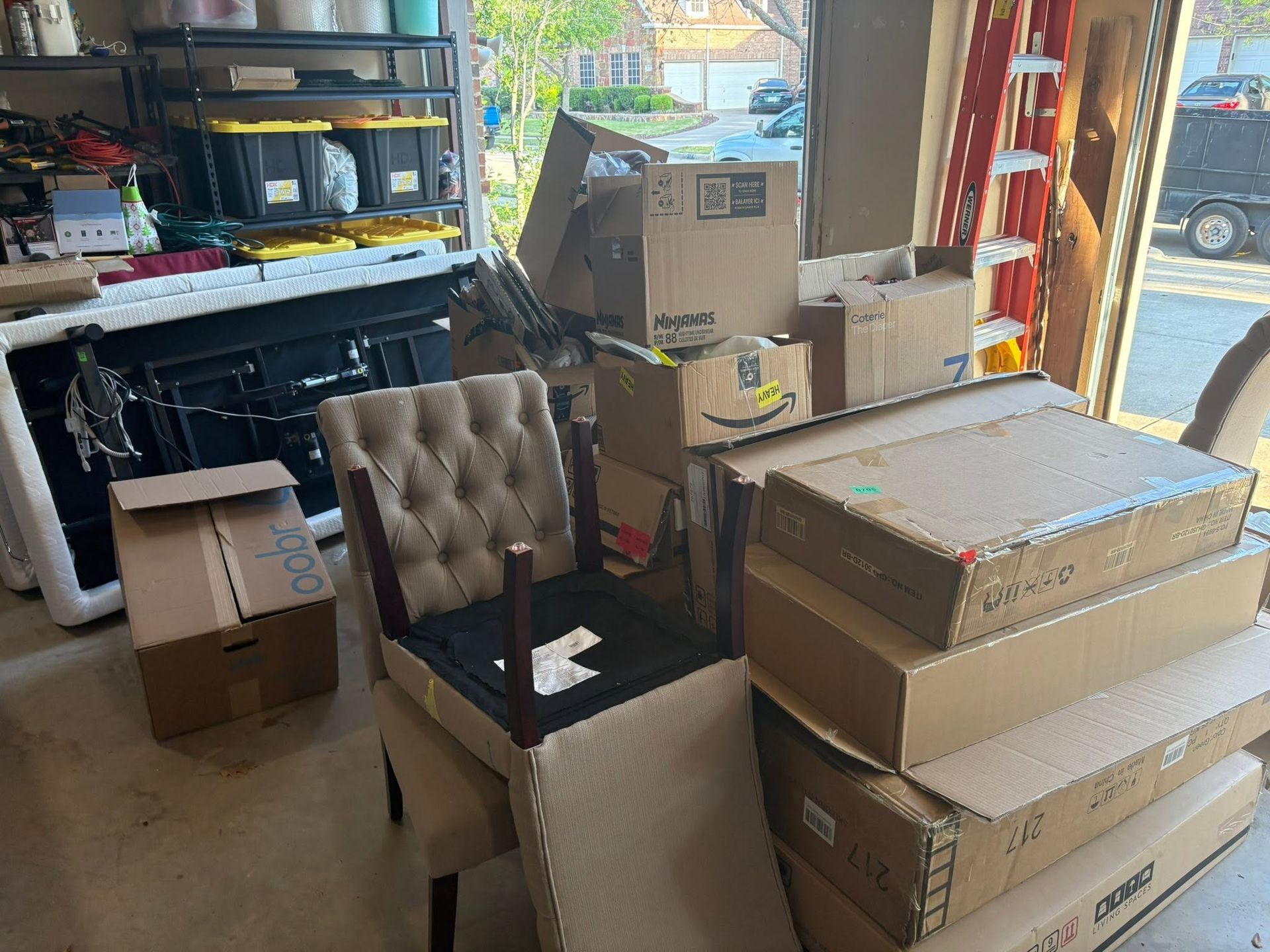 Garage cluttered with moving boxes, a black chair, and a storage shelf near an open door to a street.