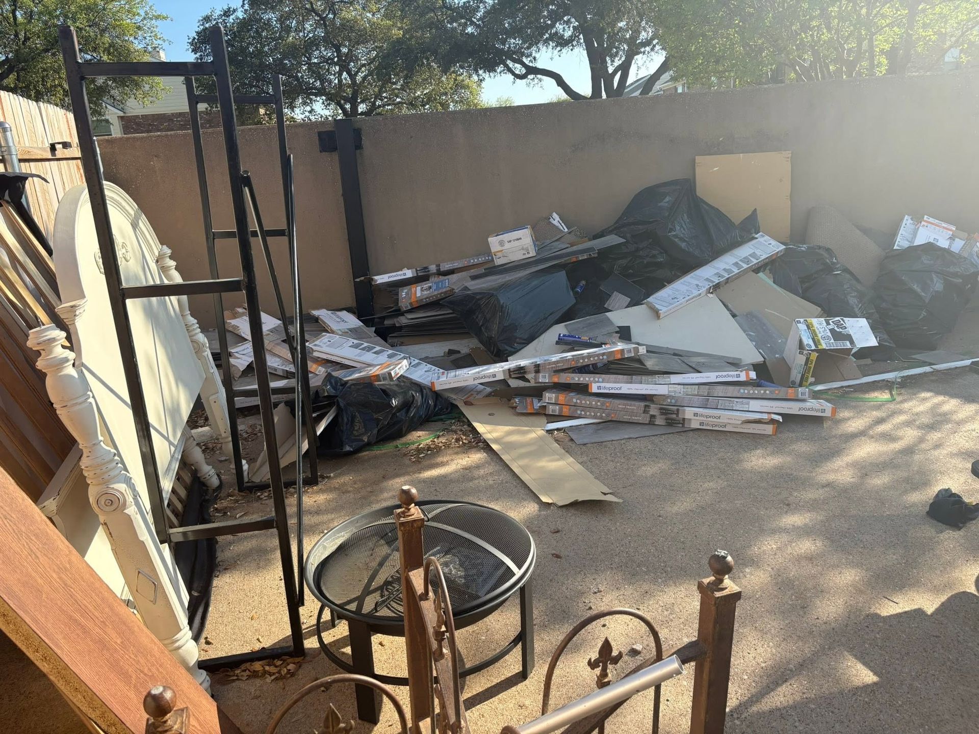 Backyard with scattered debris, broken furniture, and trash piled against a wall in sunlight.
