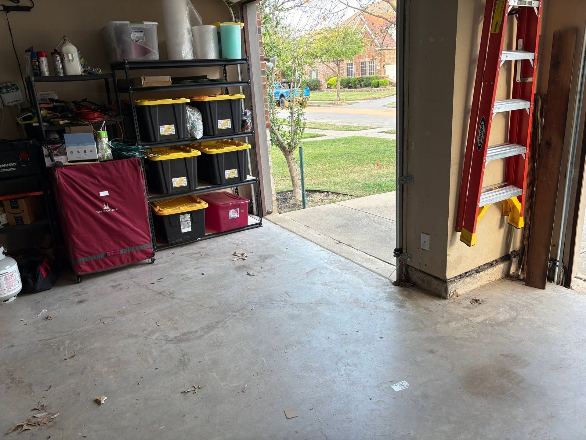 Open garage with shelves, tools, and a red ladder; driveway and yard visible through the open door.