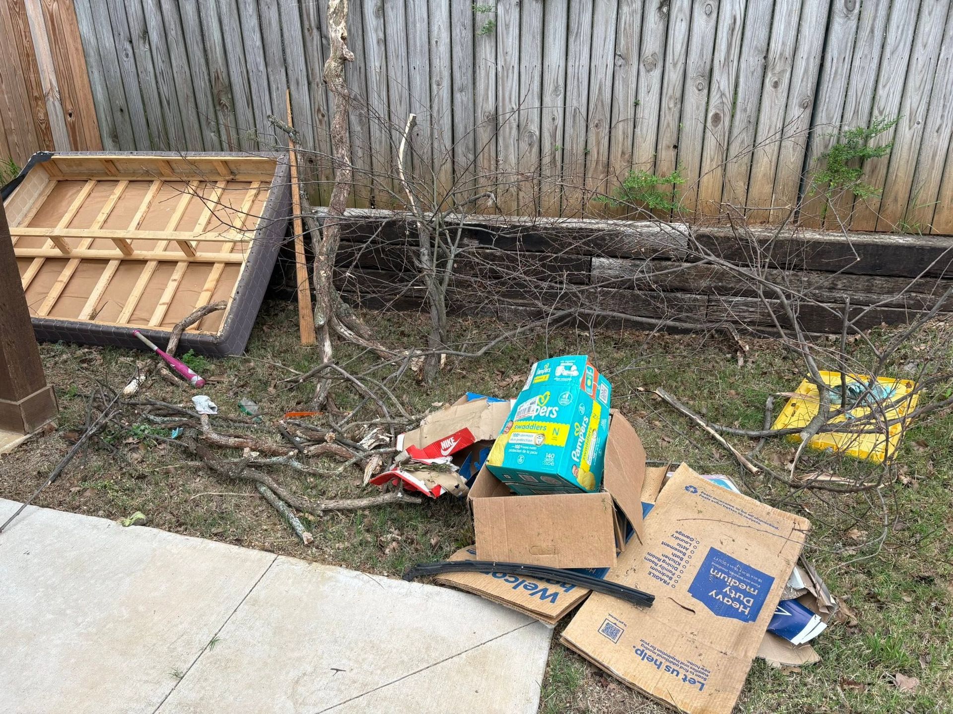 Backyard with scattered trash, cardboard boxes, and a broken wooden pallet beside a fence