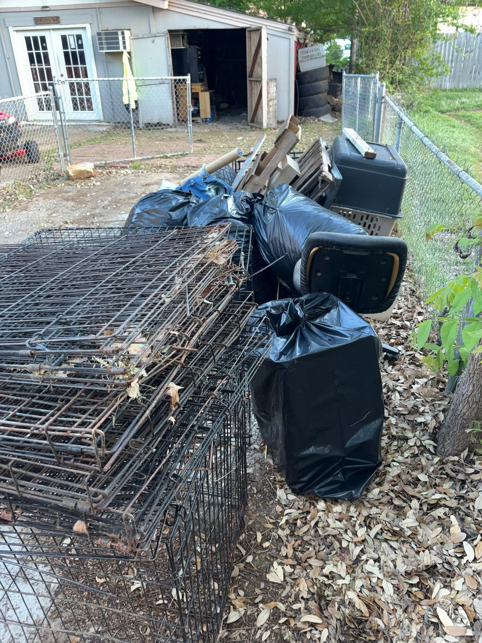 Coiled wire fencing and black bins tipped over on a dirt path beside a shed and chain-link fence.
