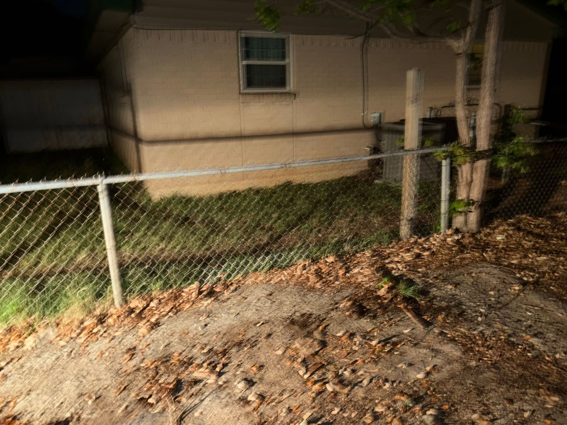 Chain-link fence beside a beige house at night, with a dirt slope and trees nearby.