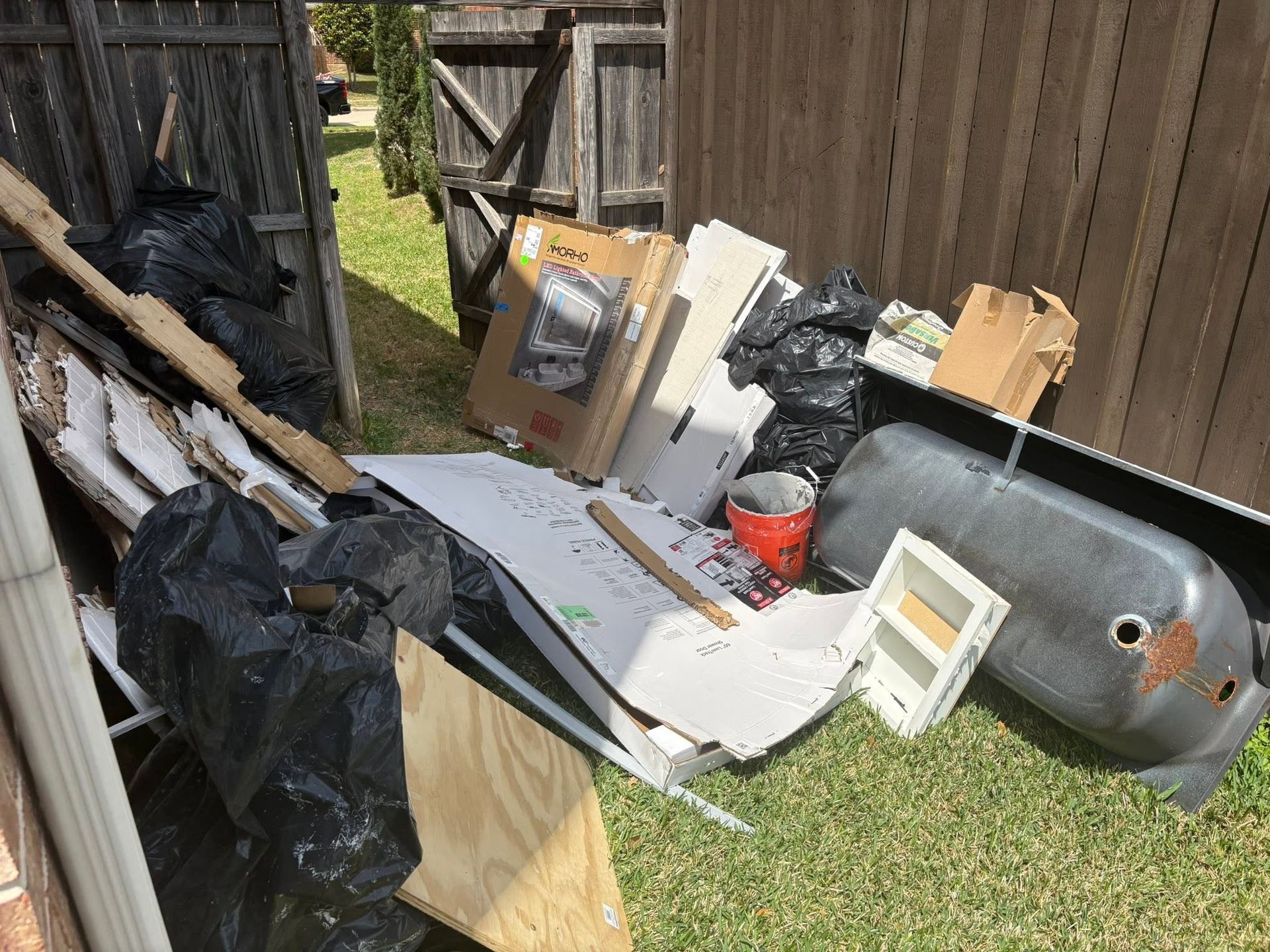 Pile of discarded cardboard, metal, and black trash bags piled beside a wooden fence on grass.