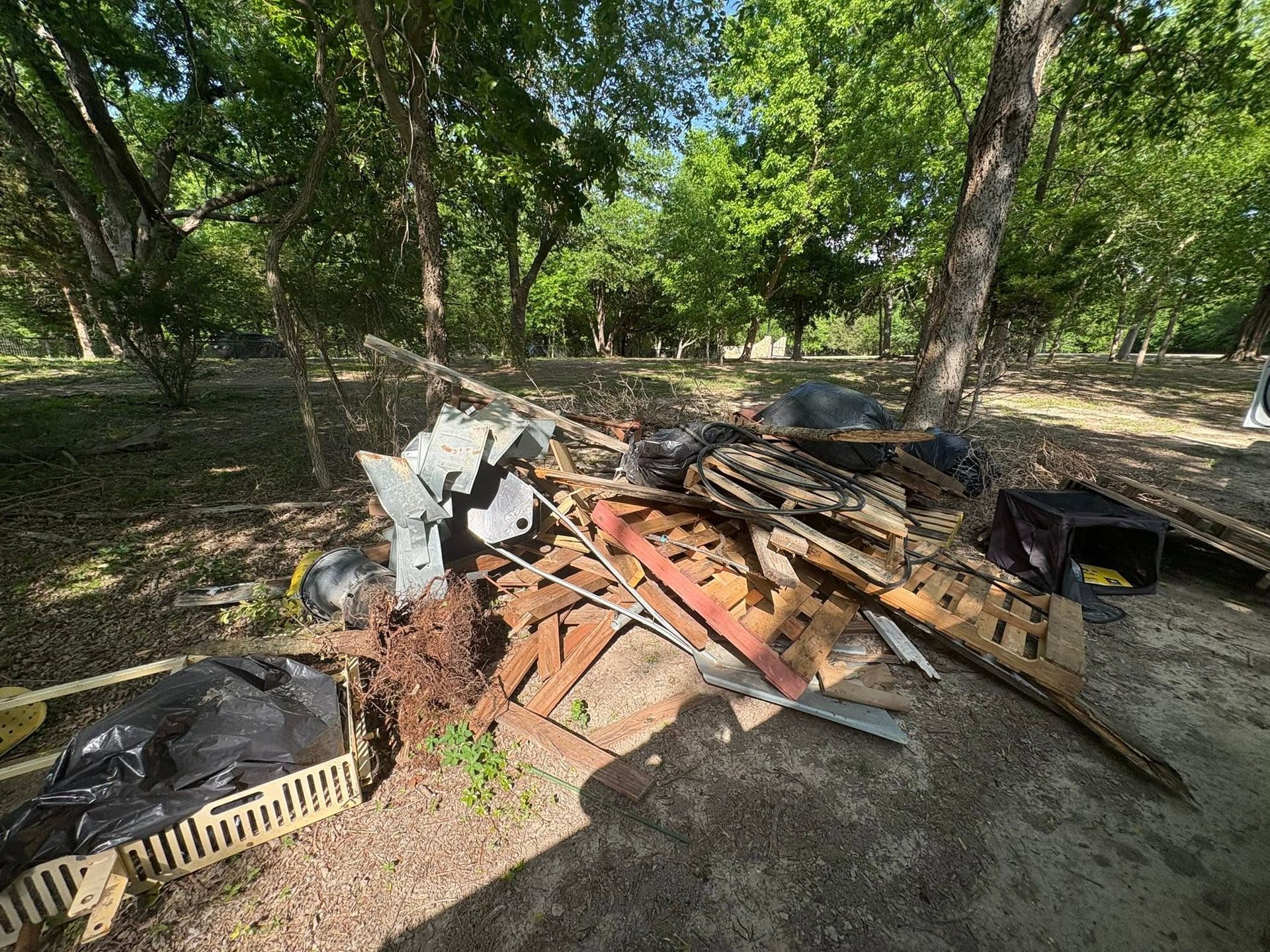 Pile of scrap wood and metal debris in a wooded outdoor area under trees.