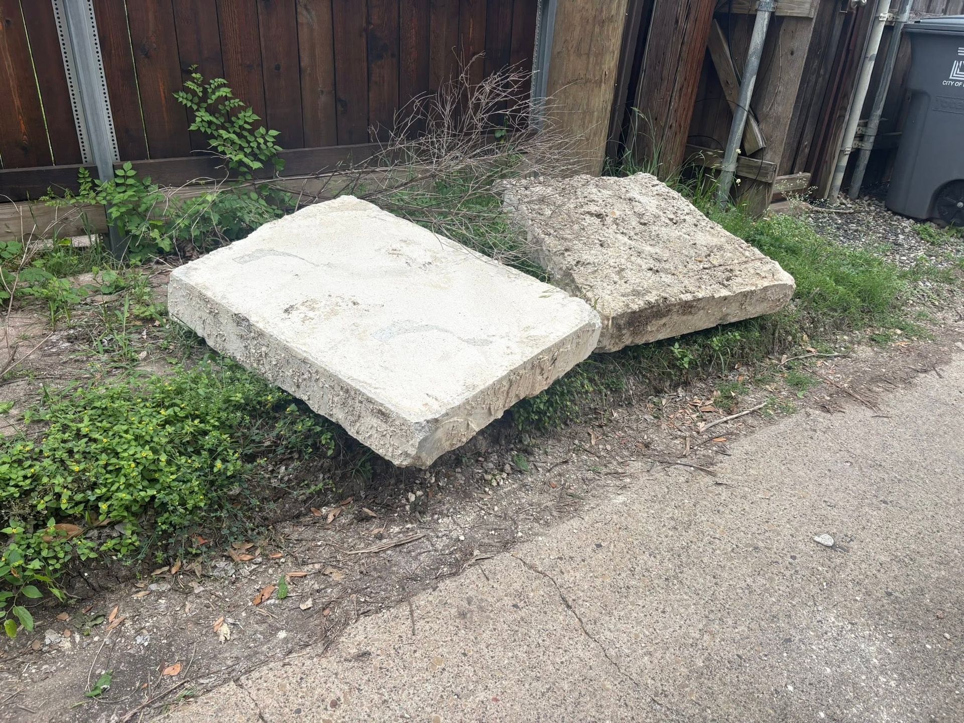 Two concrete slabs leaning by a gravel path beside a wooden fence and trash bins.