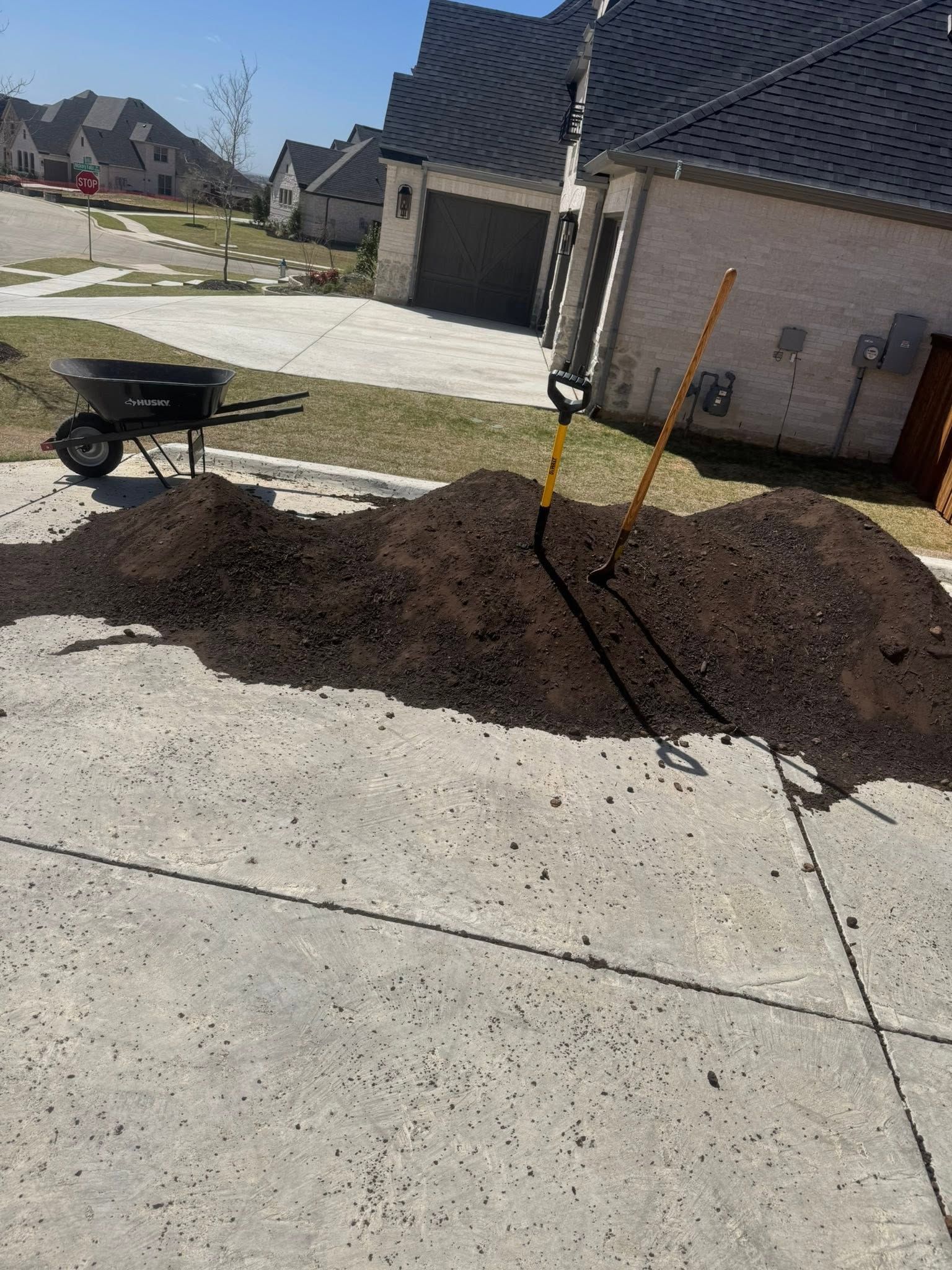 Wheelbarrow and shovel beside a large pile of mulch on a sunny driveway near a house.