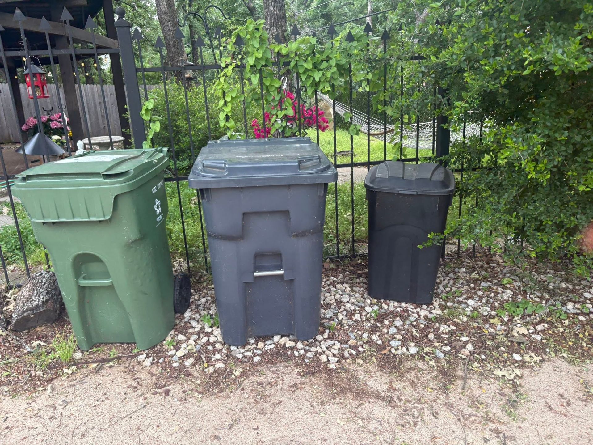 Three trash bins—green, gray, and black—lined up beside a fence in a yard.