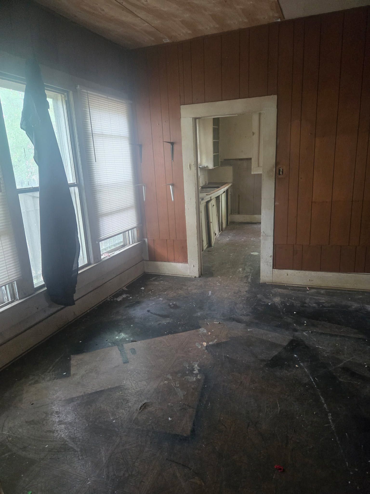 Abandoned room with torn flooring, exposed wooden walls, and doorway into a damaged kitchen area