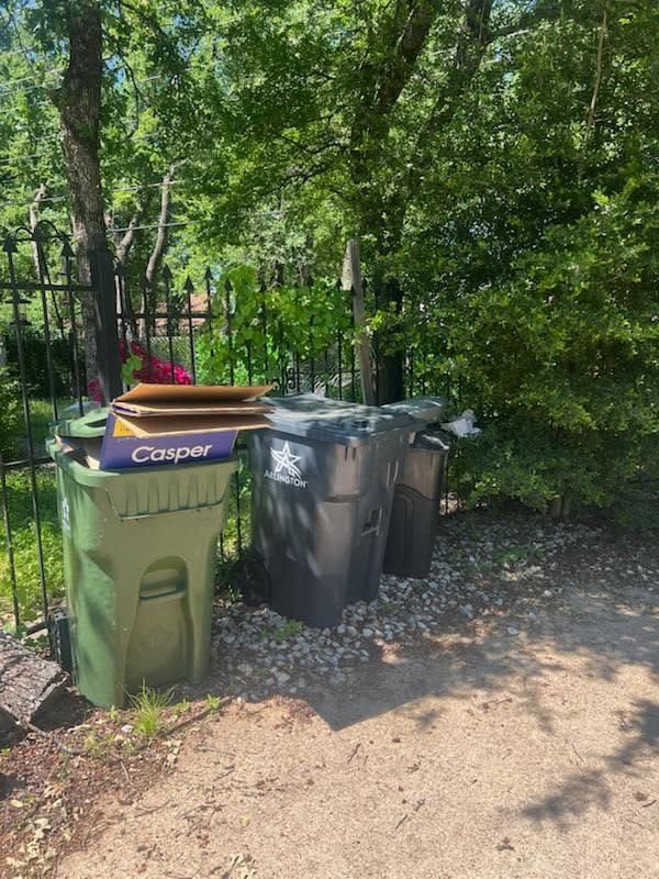 Three curbside trash bins, one green with a Casper mattress box on top, beside trees and a fence.