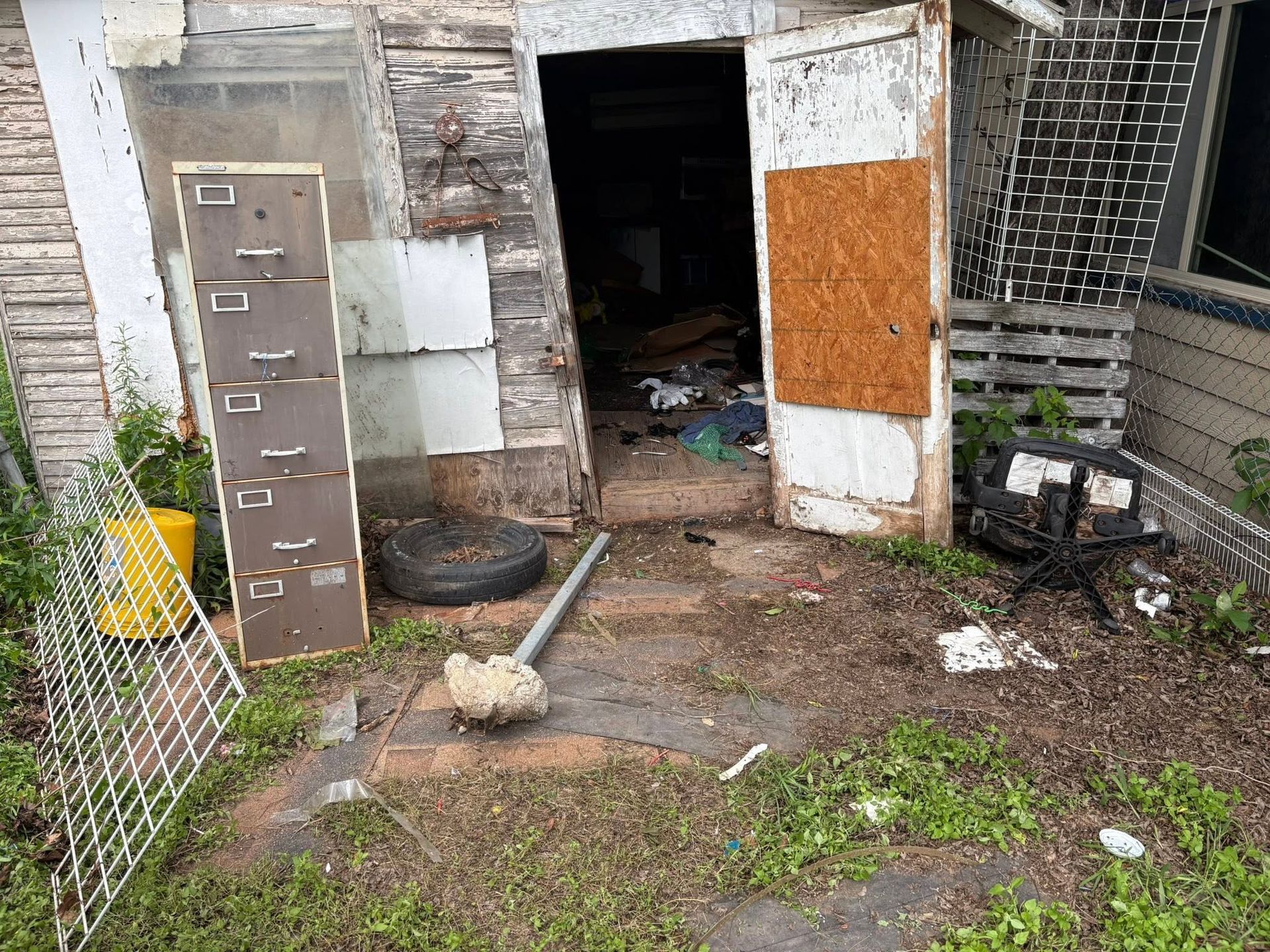 Dilapidated shed entrance with open door, debris on muddy ground, and a leaning metal cabinet beside it