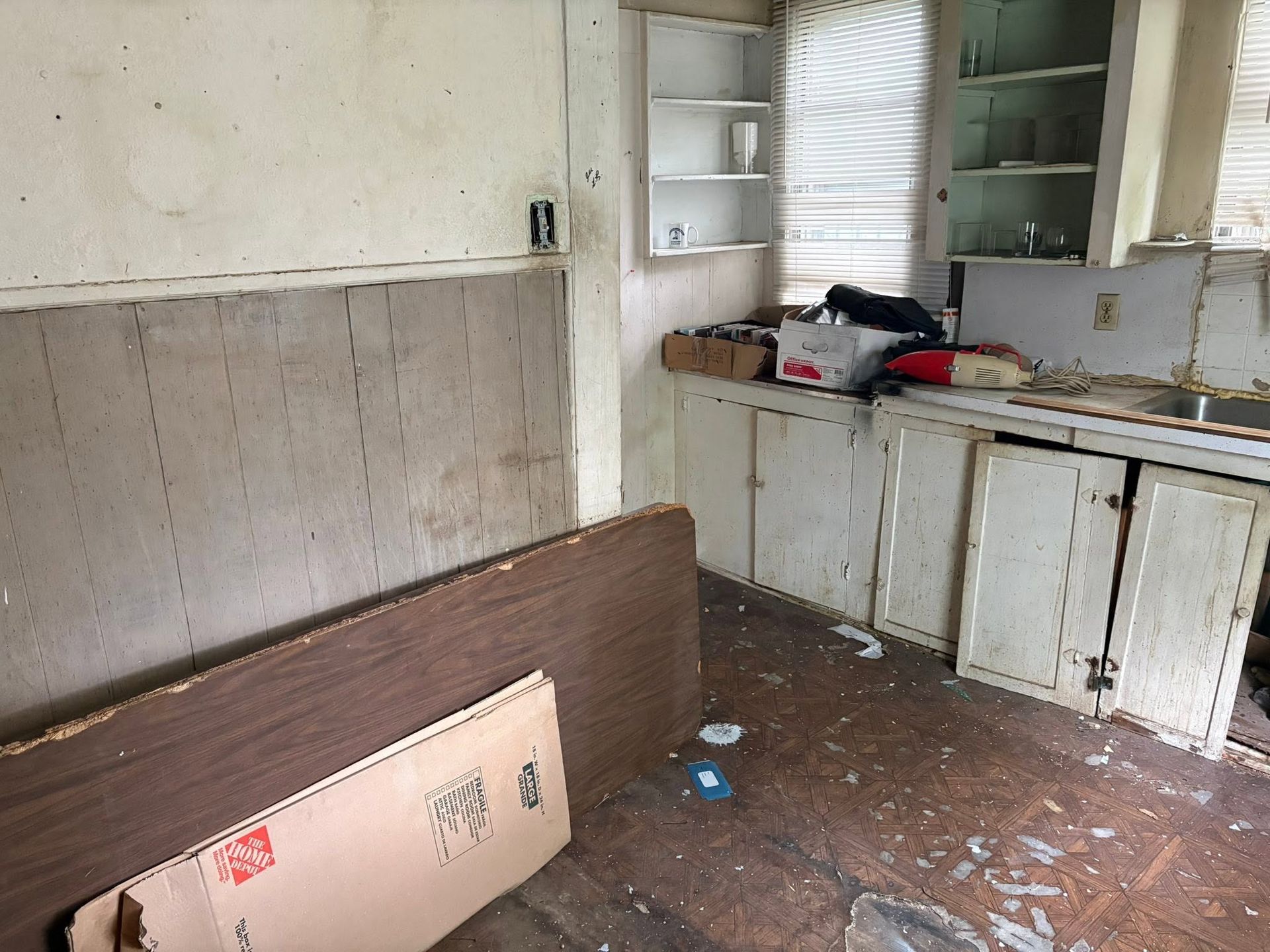 Abandoned kitchen with broken cabinets, debris on the floor, and a mattress leaning against the wall