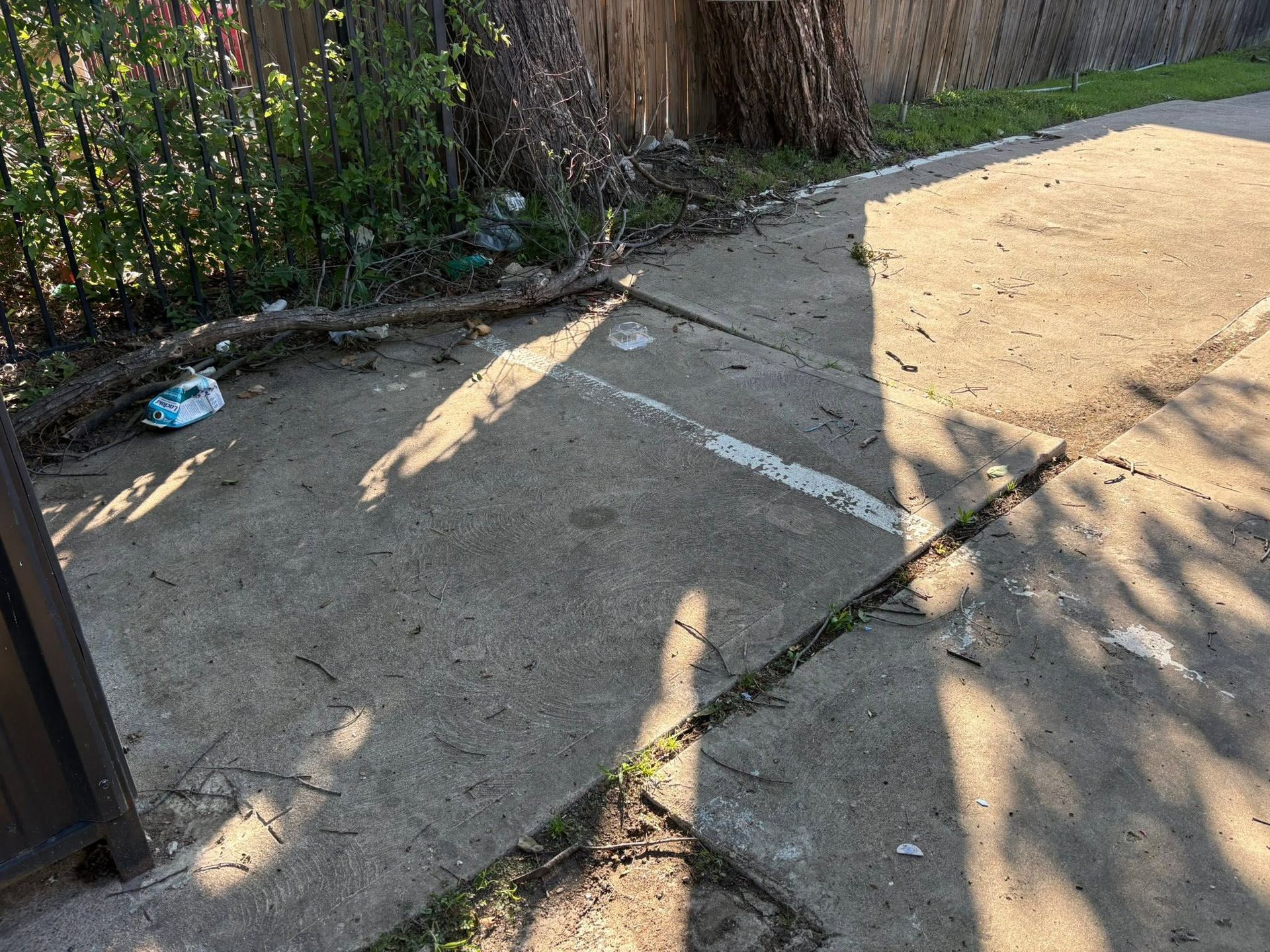 Sunlit cracked sidewalk beside a fence and tree shadows, with a concrete ramp or curb cut.
