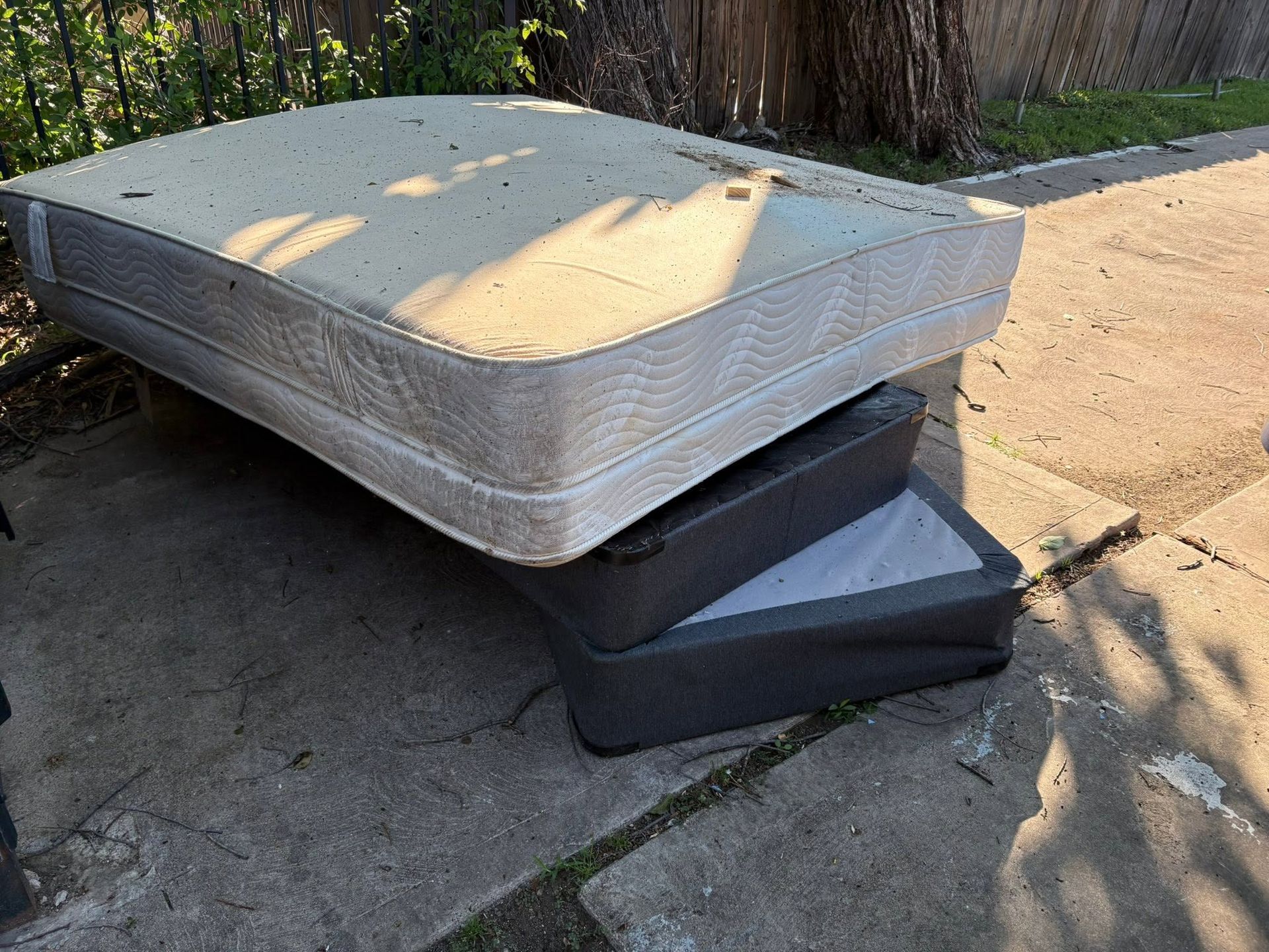Two stacked mattresses on a concrete patio, one gray and one black, in sunlight.