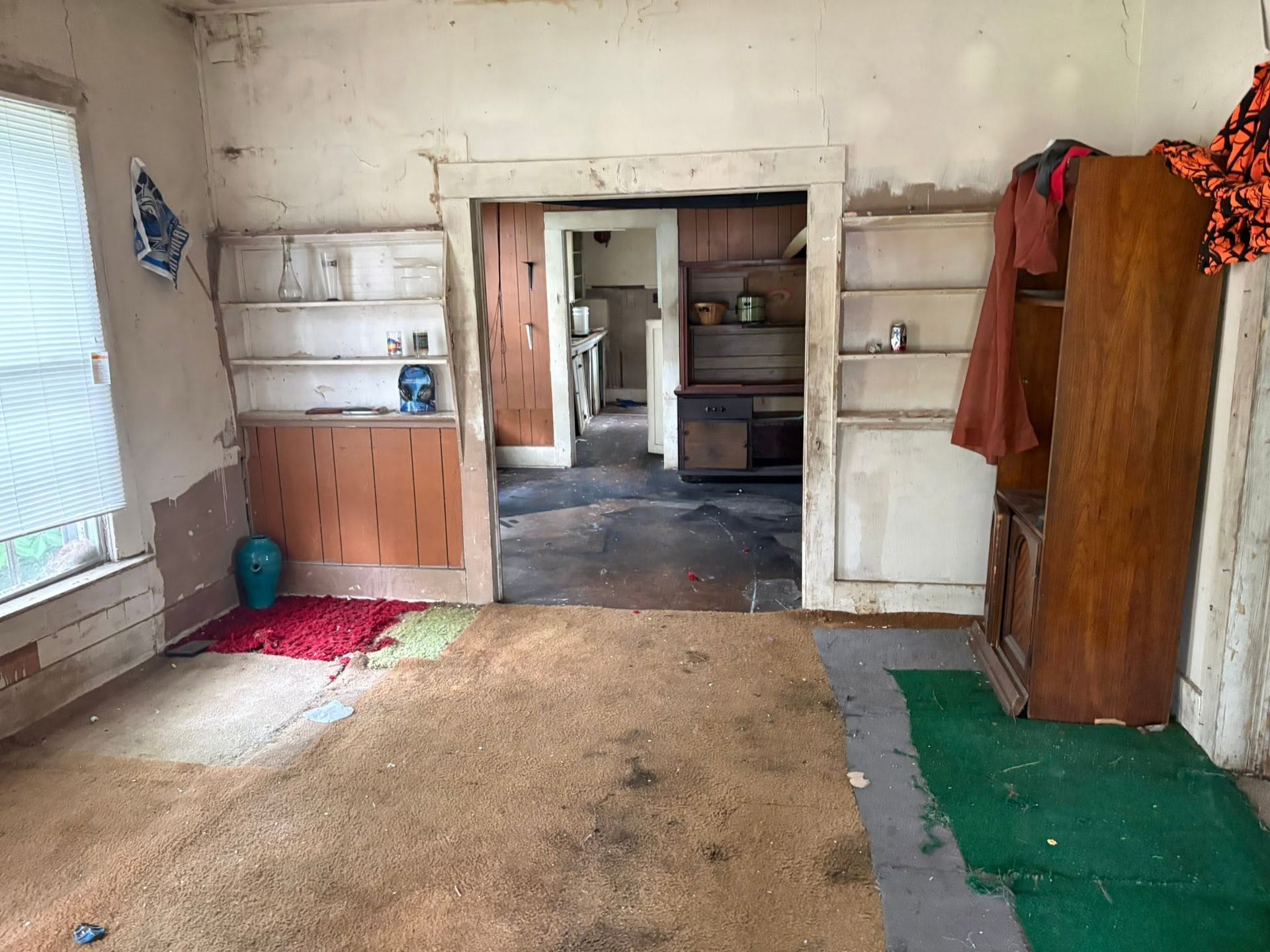 Sparse, worn room with peeling walls, a doorway to another room, and colorful mats on the floor