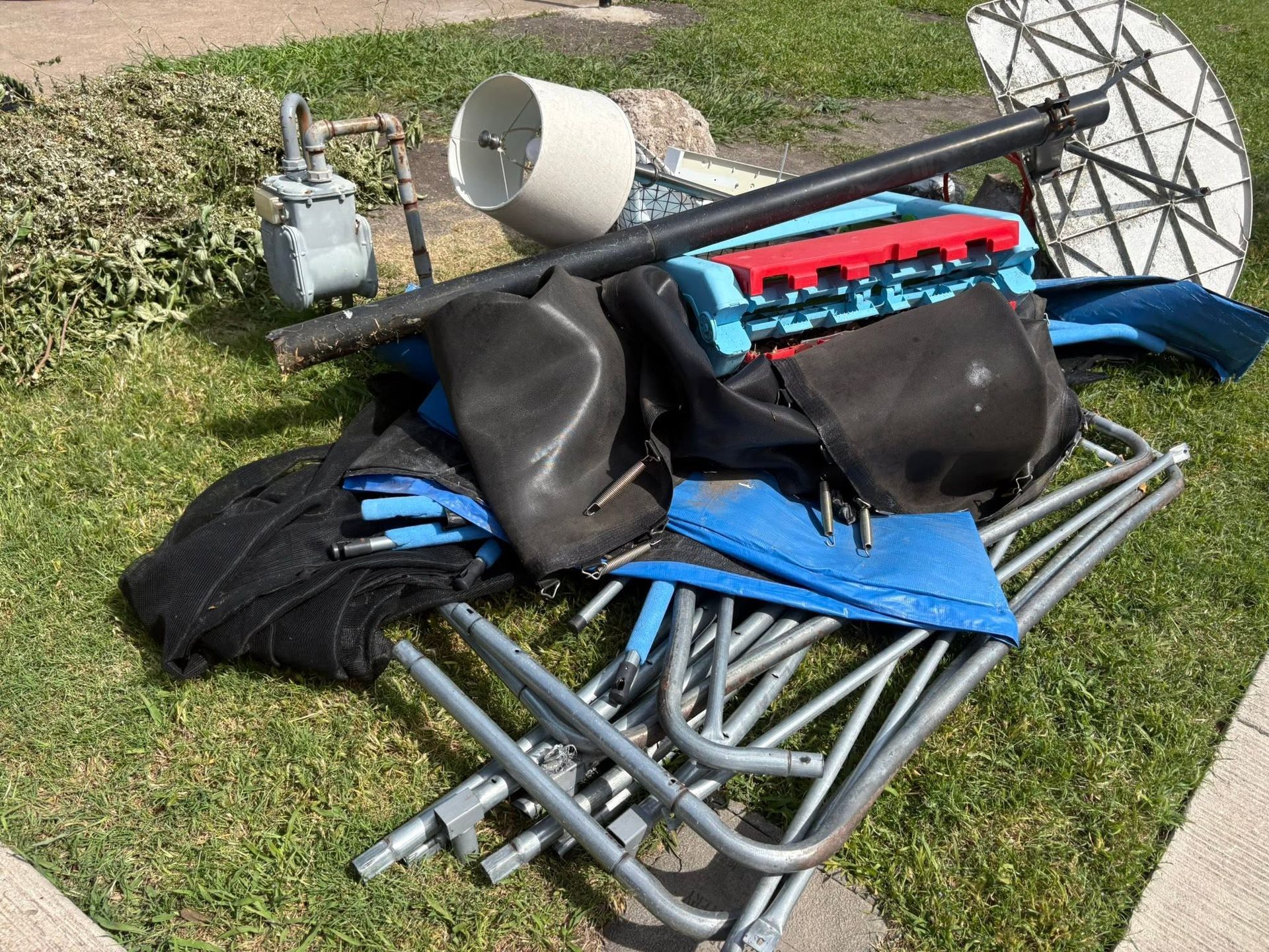 Folded camping chairs, bags, and a satellite dish piled on grass beside a sidewalk.