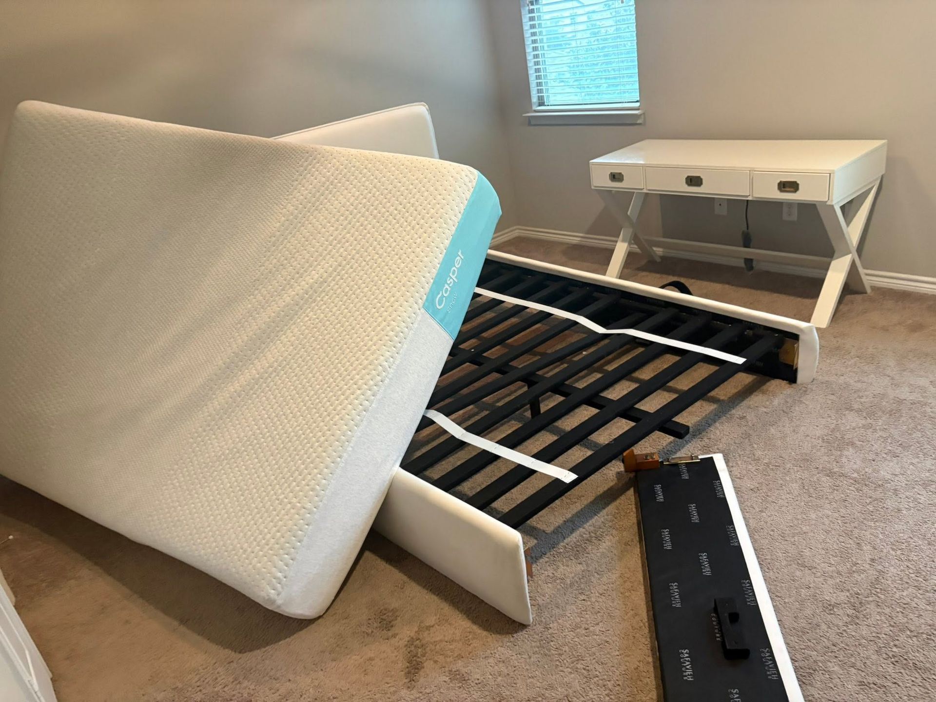 Mattress and bed frame parts scattered on a bedroom floor beside a white nightstand