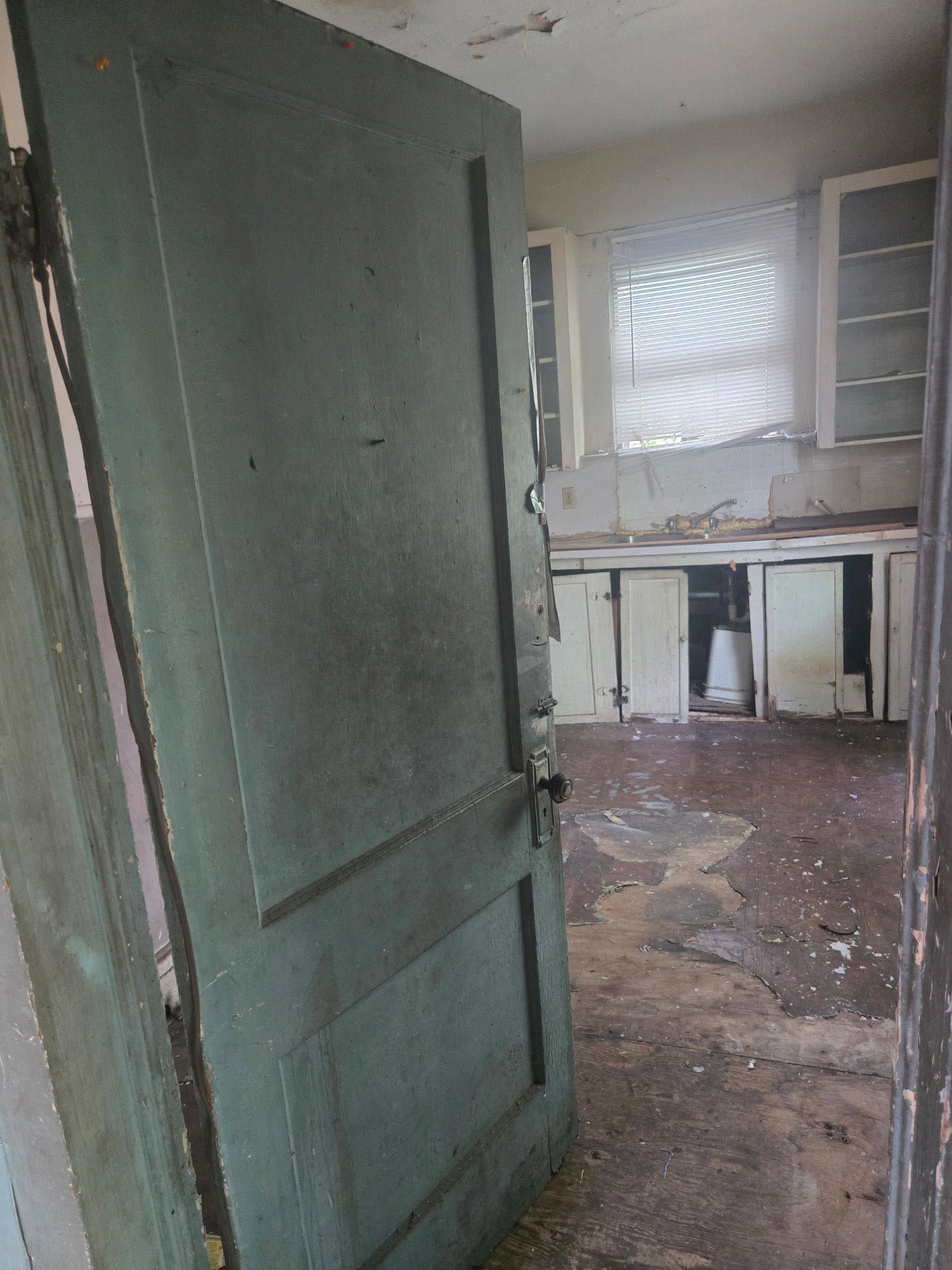 Open green door leading to a dusty, damaged room with a window and cabinets.