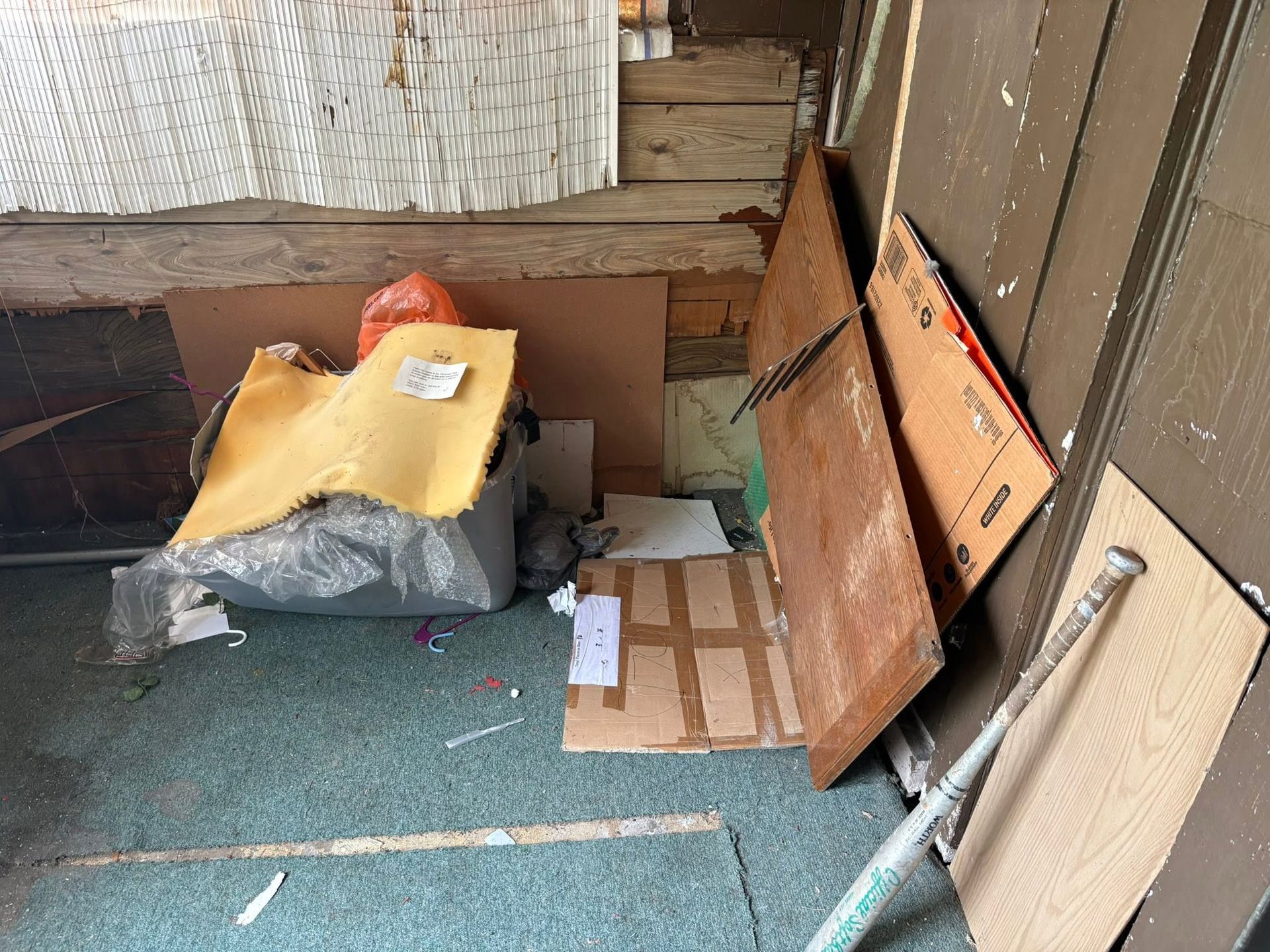 Small cluttered porch with cardboard boxes, trash bags, and wood leaning against the wall.