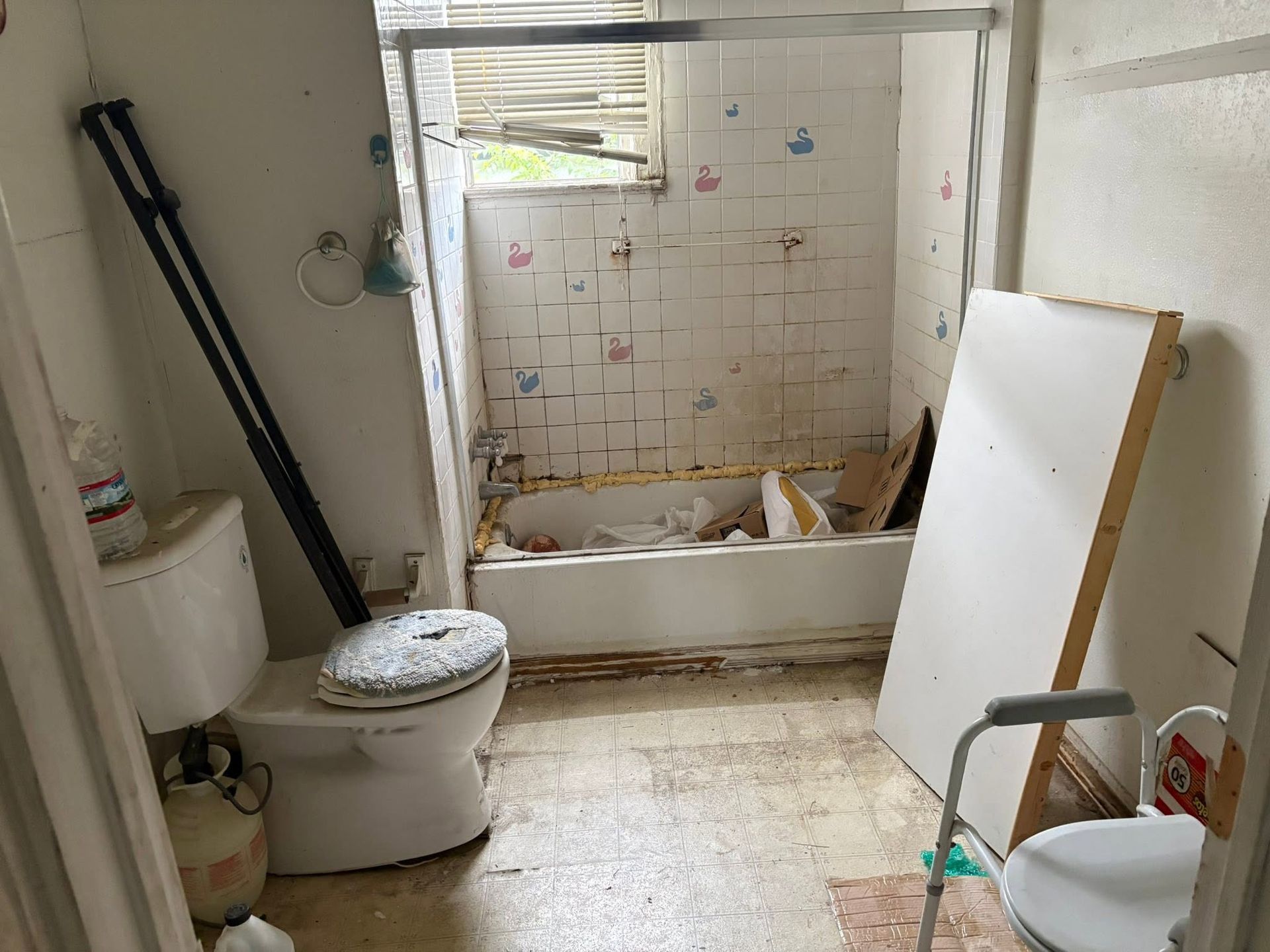 Cluttered bathroom with a toilet, bathtub, sink, and a white board leaning by the tub.