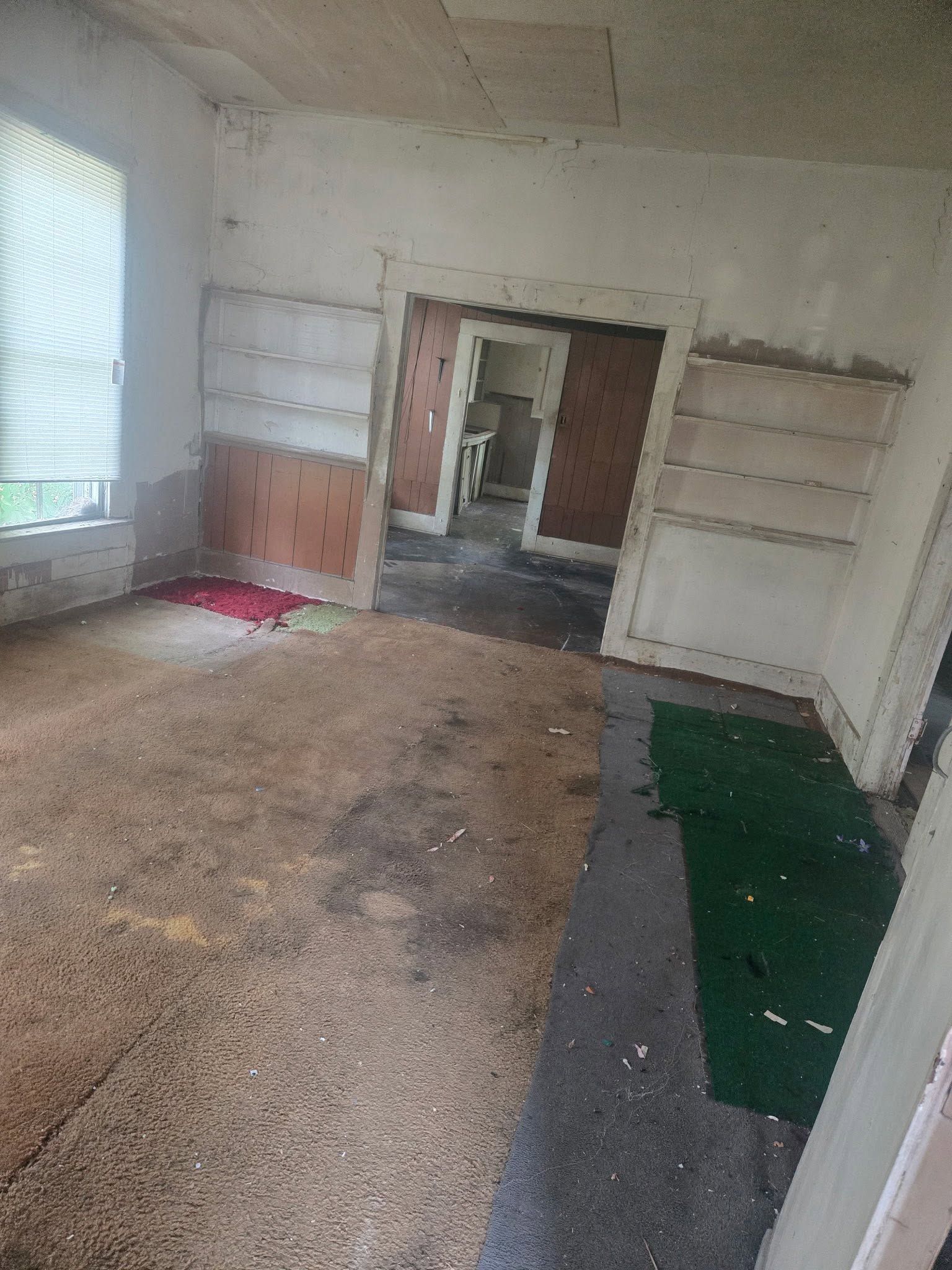 Empty, worn room with peeling walls, a window left, and doorway to another room.