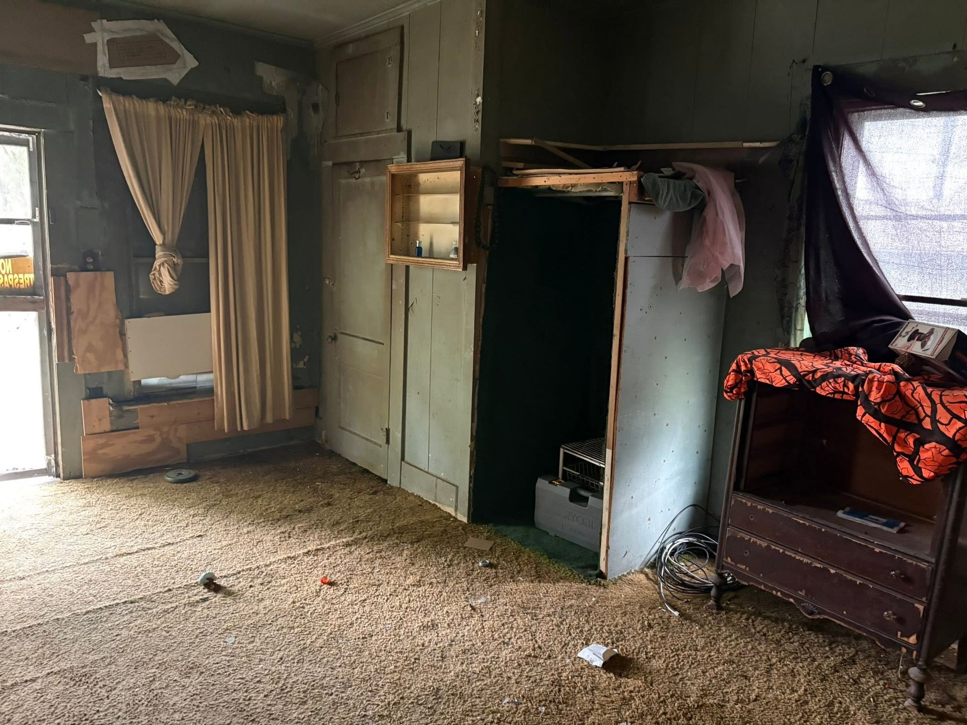 Sparse room with worn carpet, curtained window, open cabinet, and bed covered in red blanket