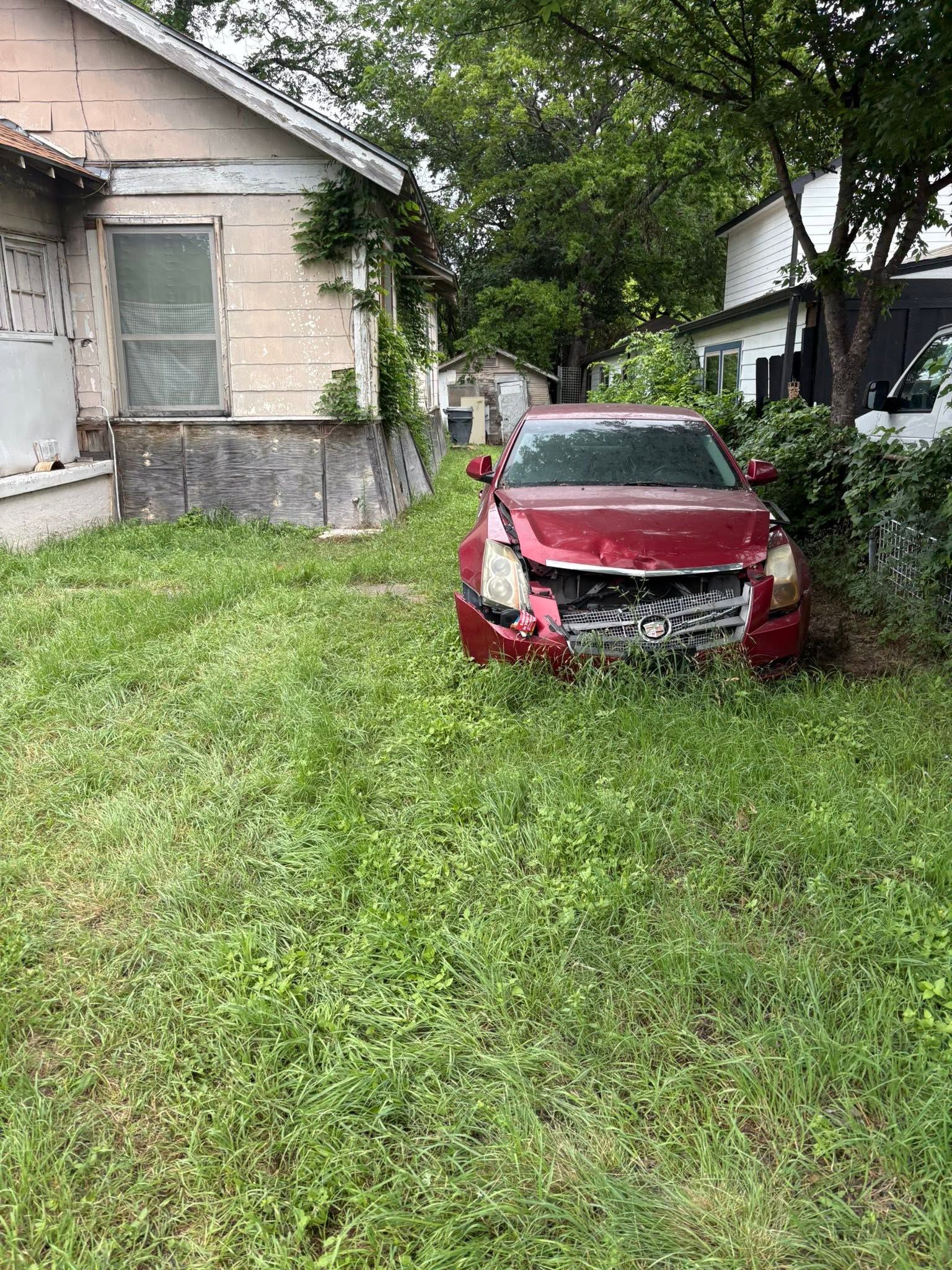 Red car parked in tall grass beside a house and trees