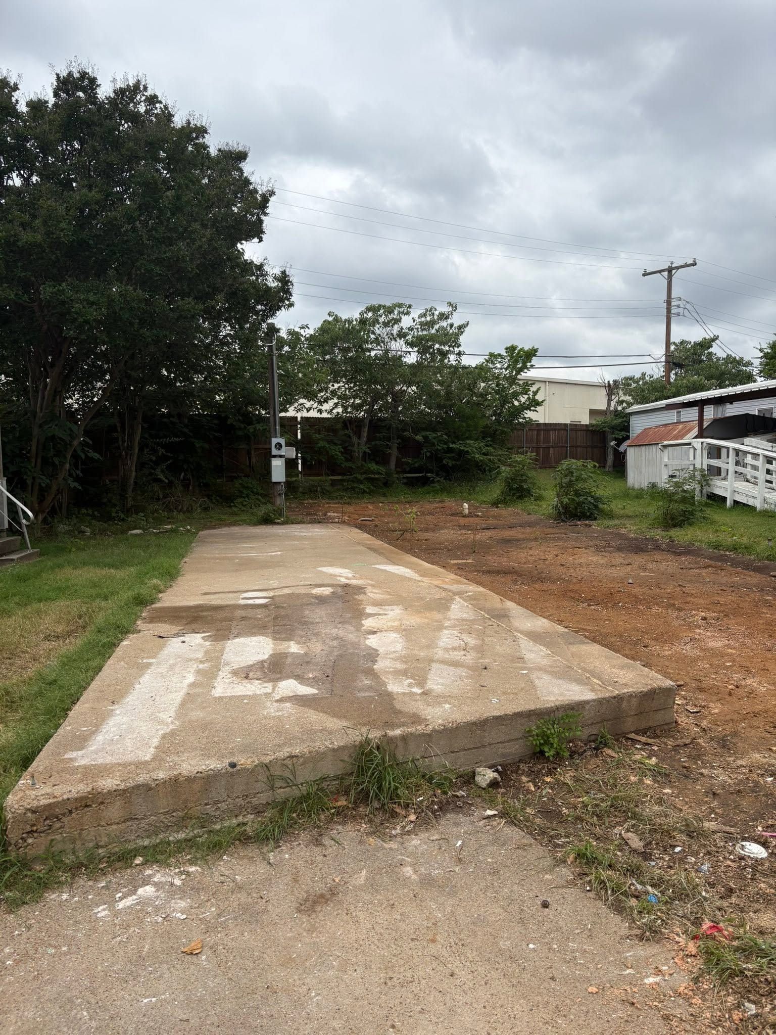 Empty concrete slab beside a dirt road, with trees, houses, and a utility pole in the background