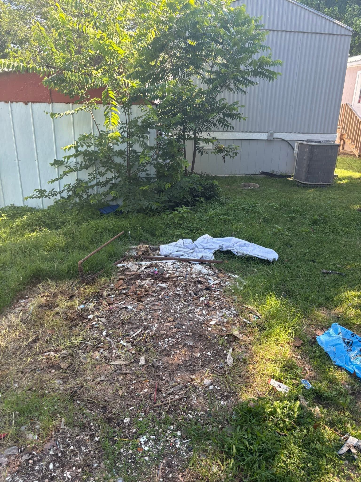 Backyard with a compost pile, tree, grass, and a small shed or fence in the background