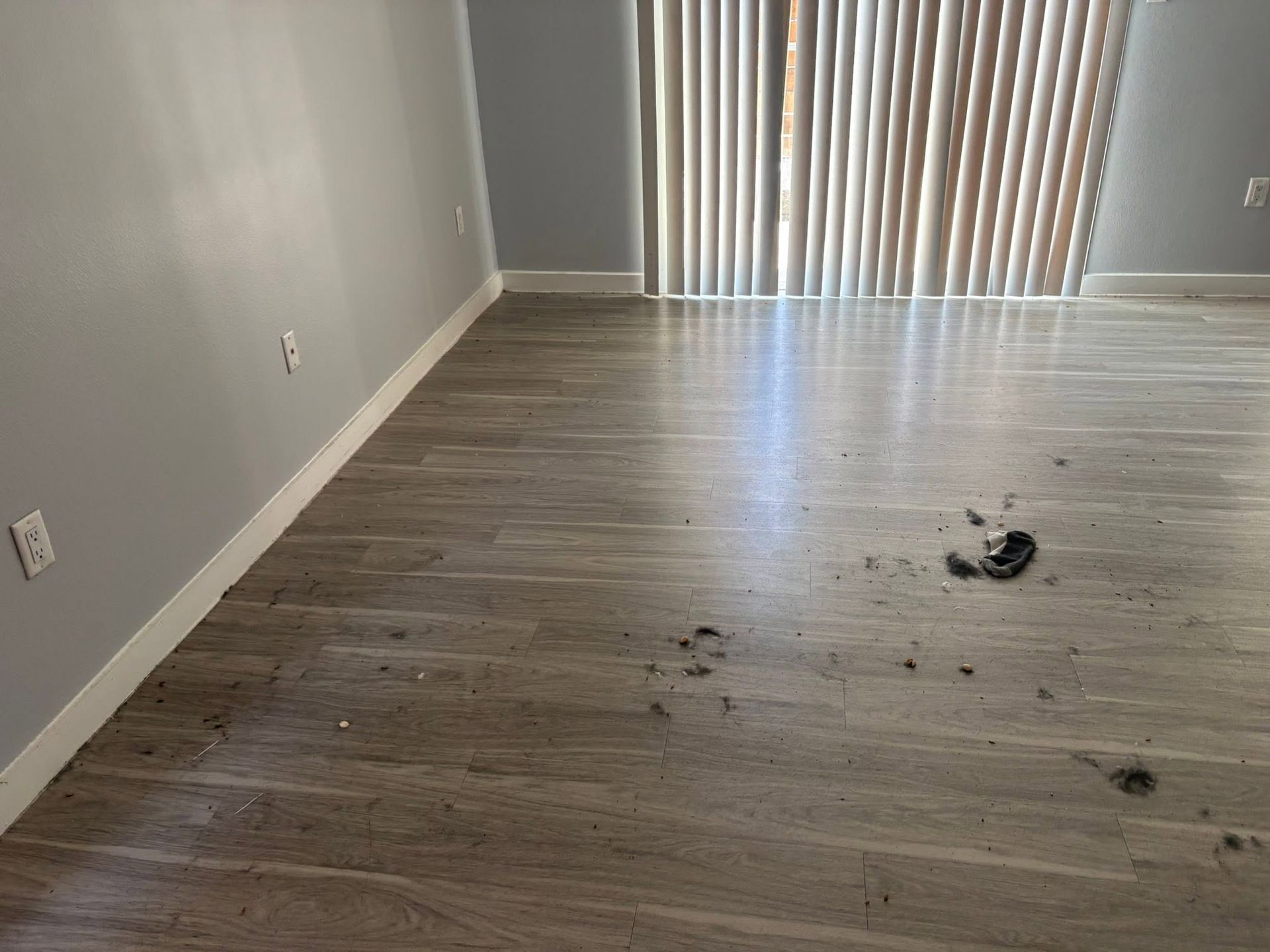 Empty room with gray walls, wood floor, and scattered debris near the sliding glass door.