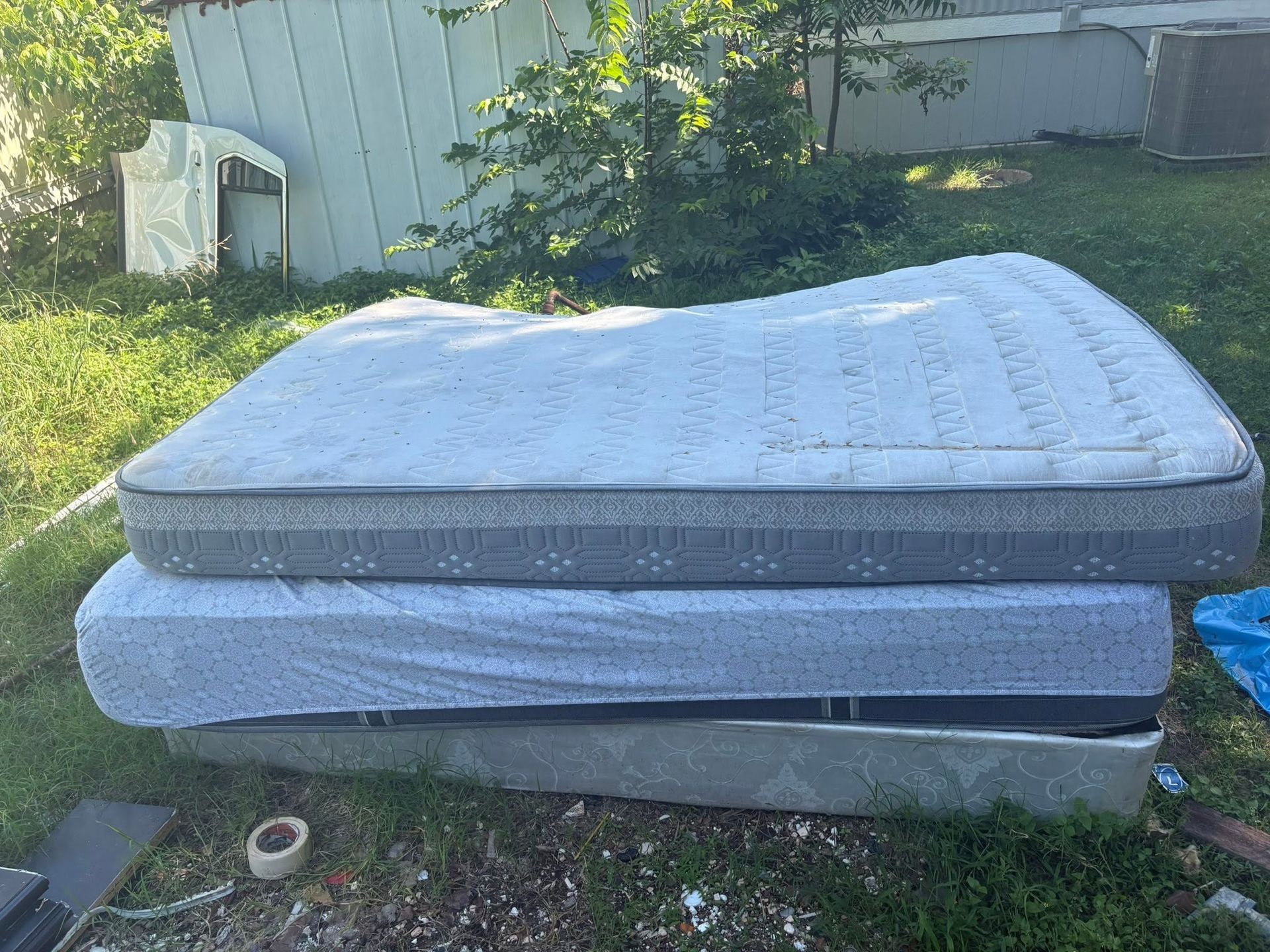 Two mattresses stacked outdoors in a grassy yard near a fence