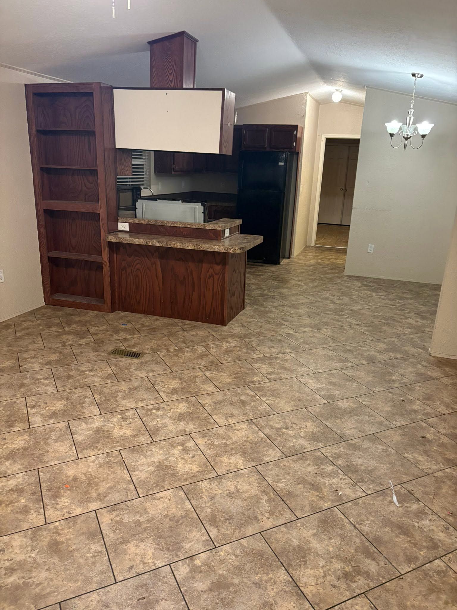 Empty kitchen with tiled floor, wood cabinets, and a chandelier in a large open room.