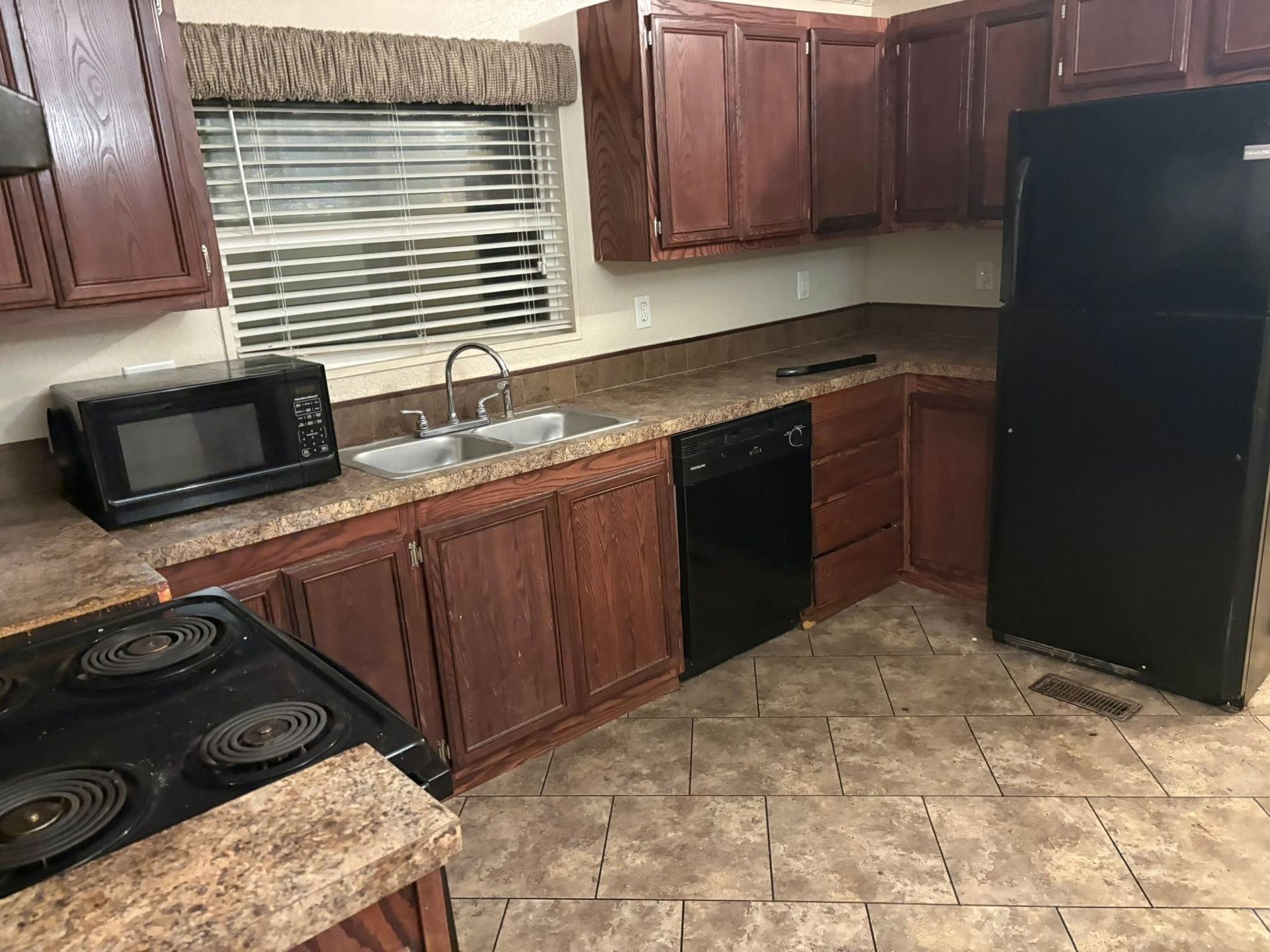 Empty kitchen with dark wood cabinets, black appliances, tiled floor, and a window above the sink.