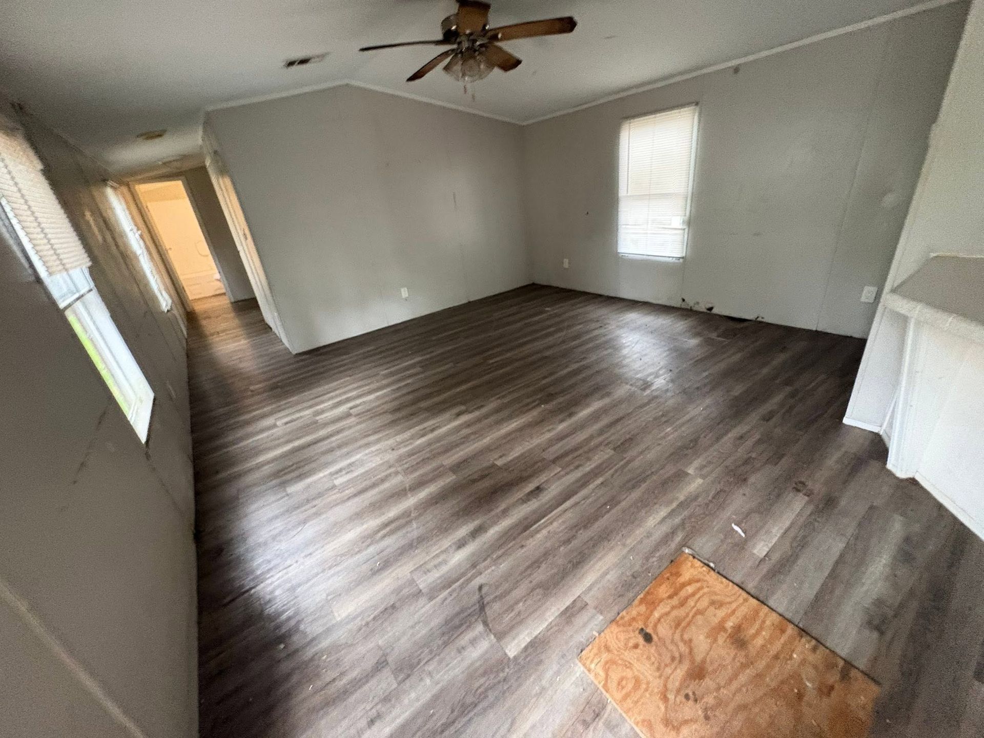Empty living room with gray walls, wood-look flooring, ceiling fan, and sunlight from windows