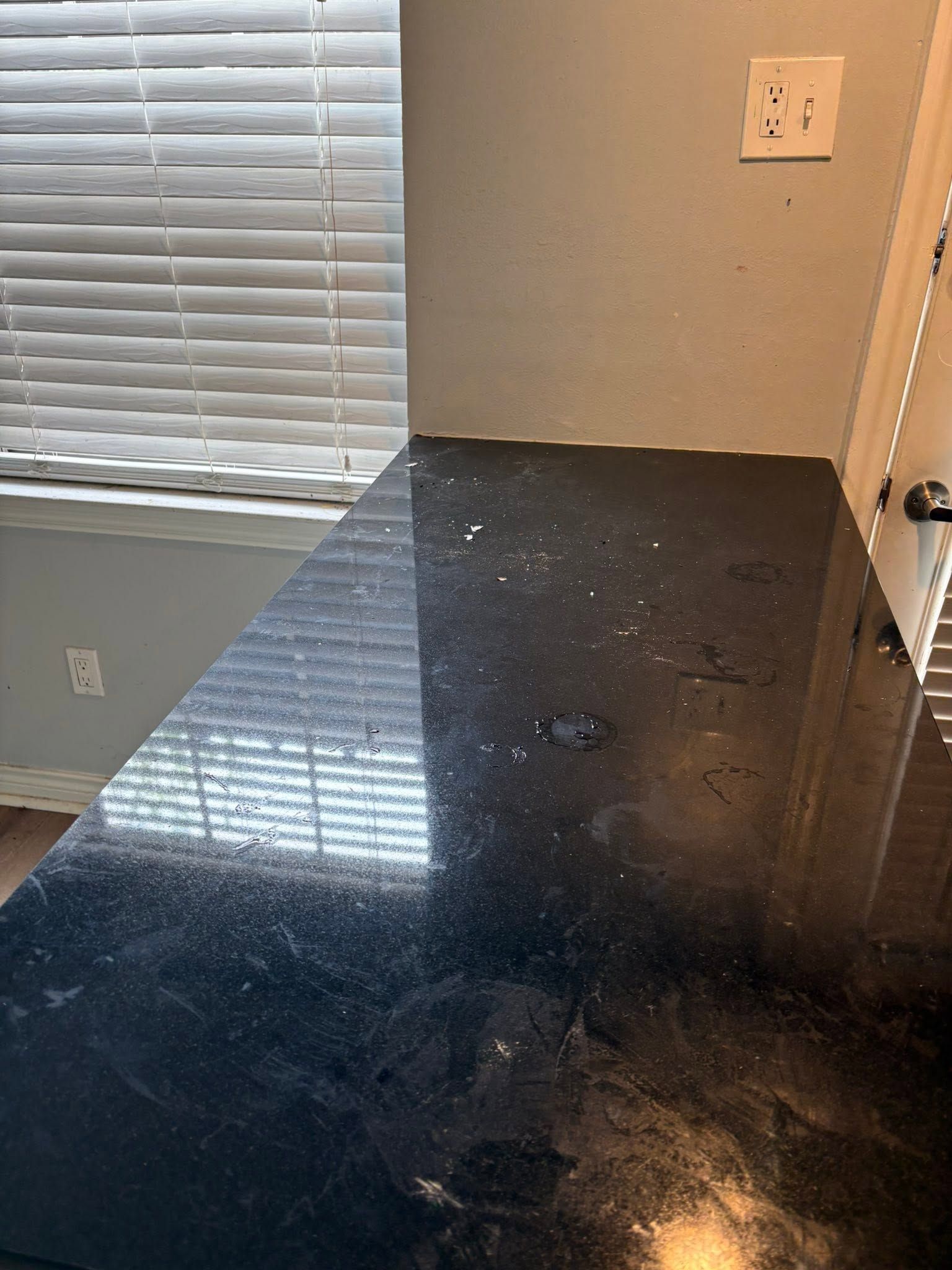 Black granite countertop beside a window with blinds and a wall outlet.