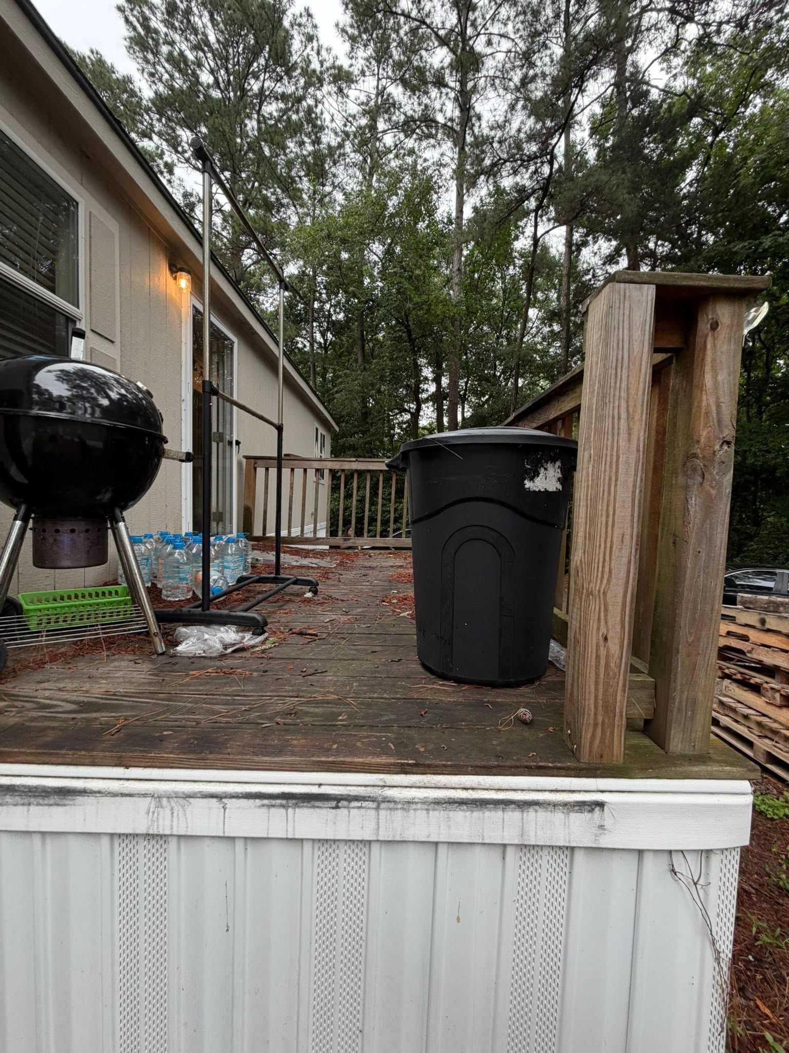 Backyard deck with grill, black trash bin, and wooden post beside a house and trees