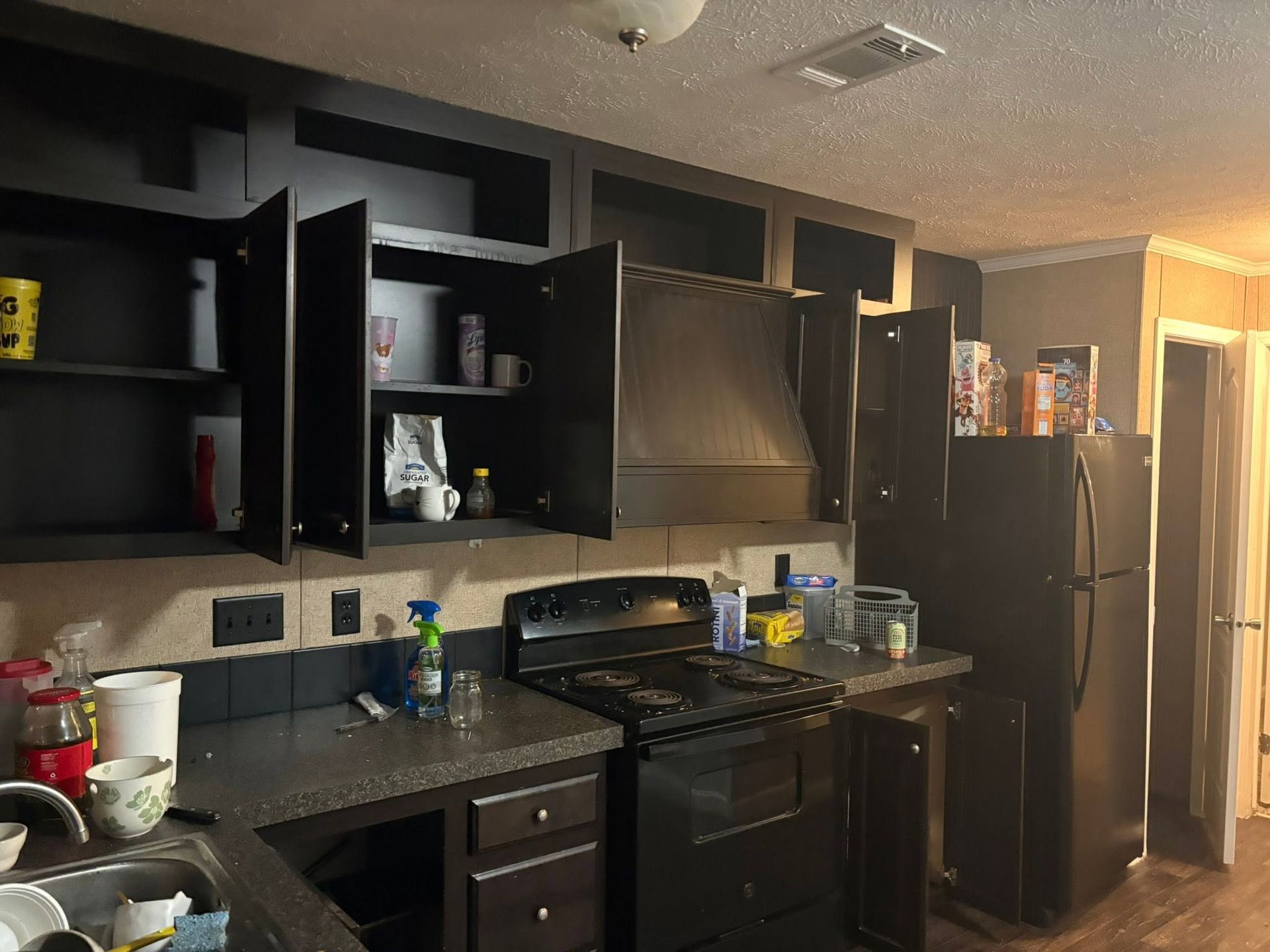 Dark kitchen with black cabinets, stove, sink, and refrigerator under warm light.