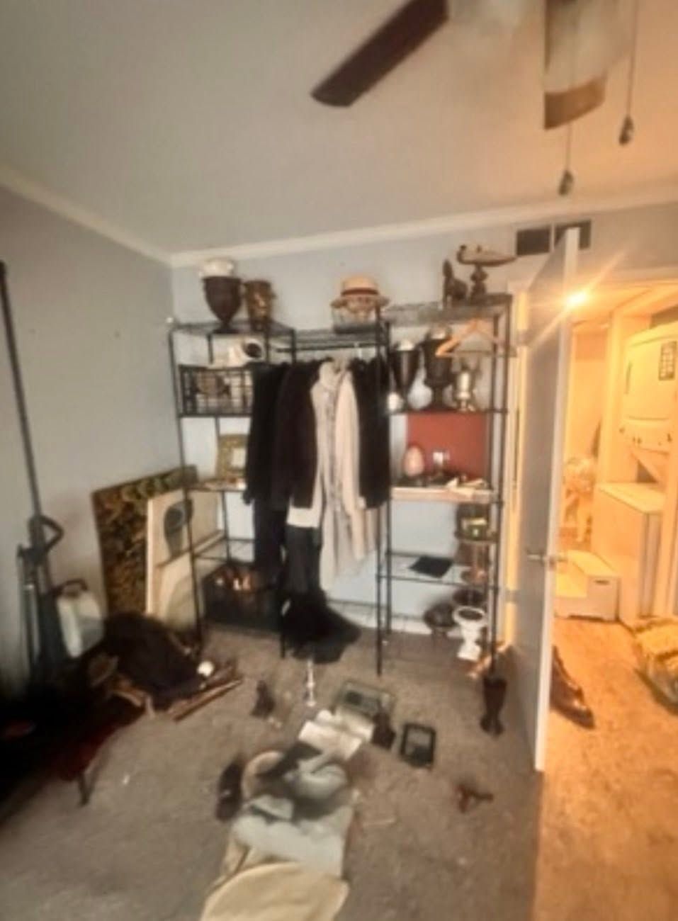 Cluttered bedroom with black clothing rack, shelves, scattered items, and an open doorway to a lit hall