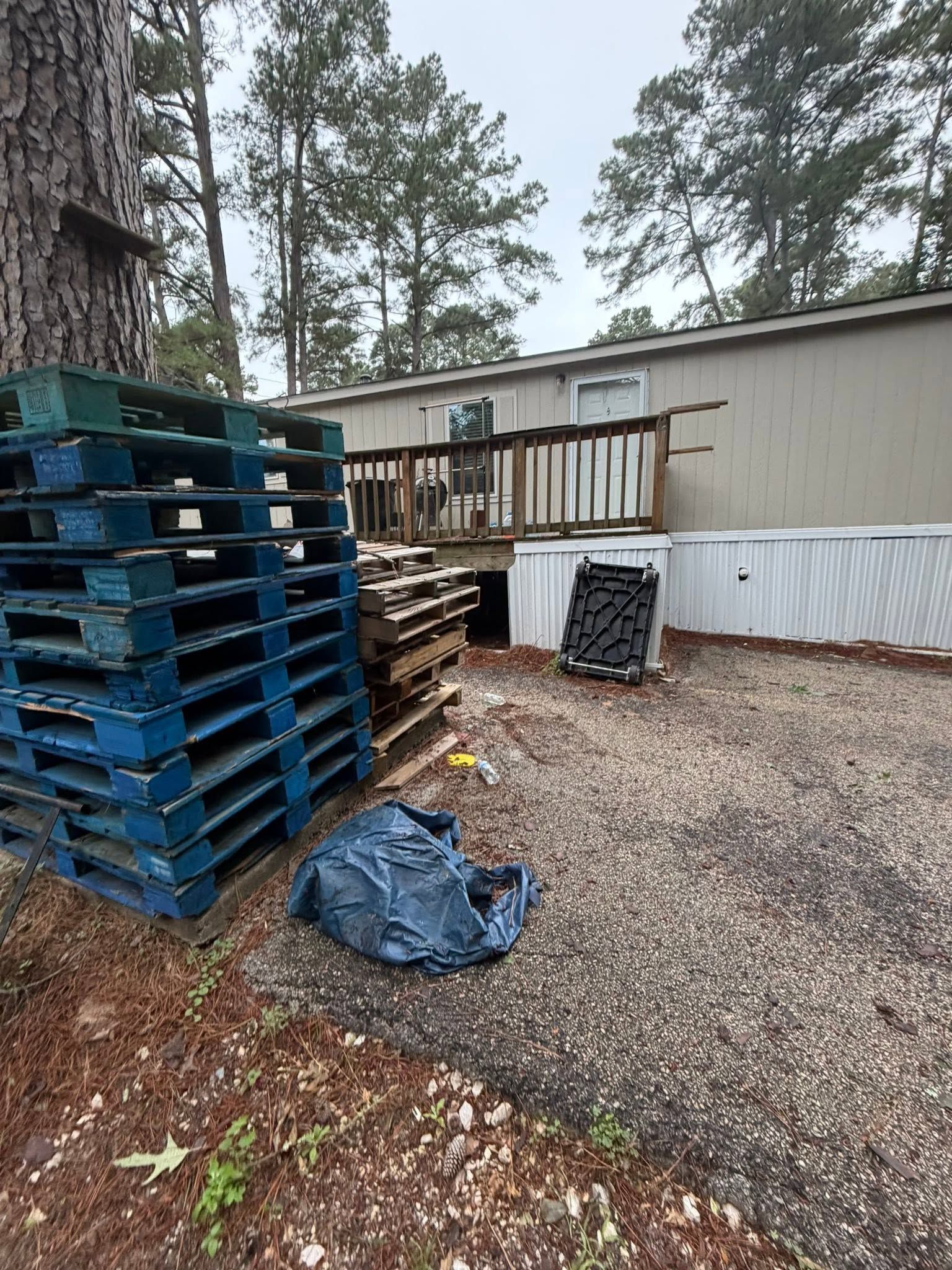 Stacked blue pallets beside a mobile home in a wooded gravel yard