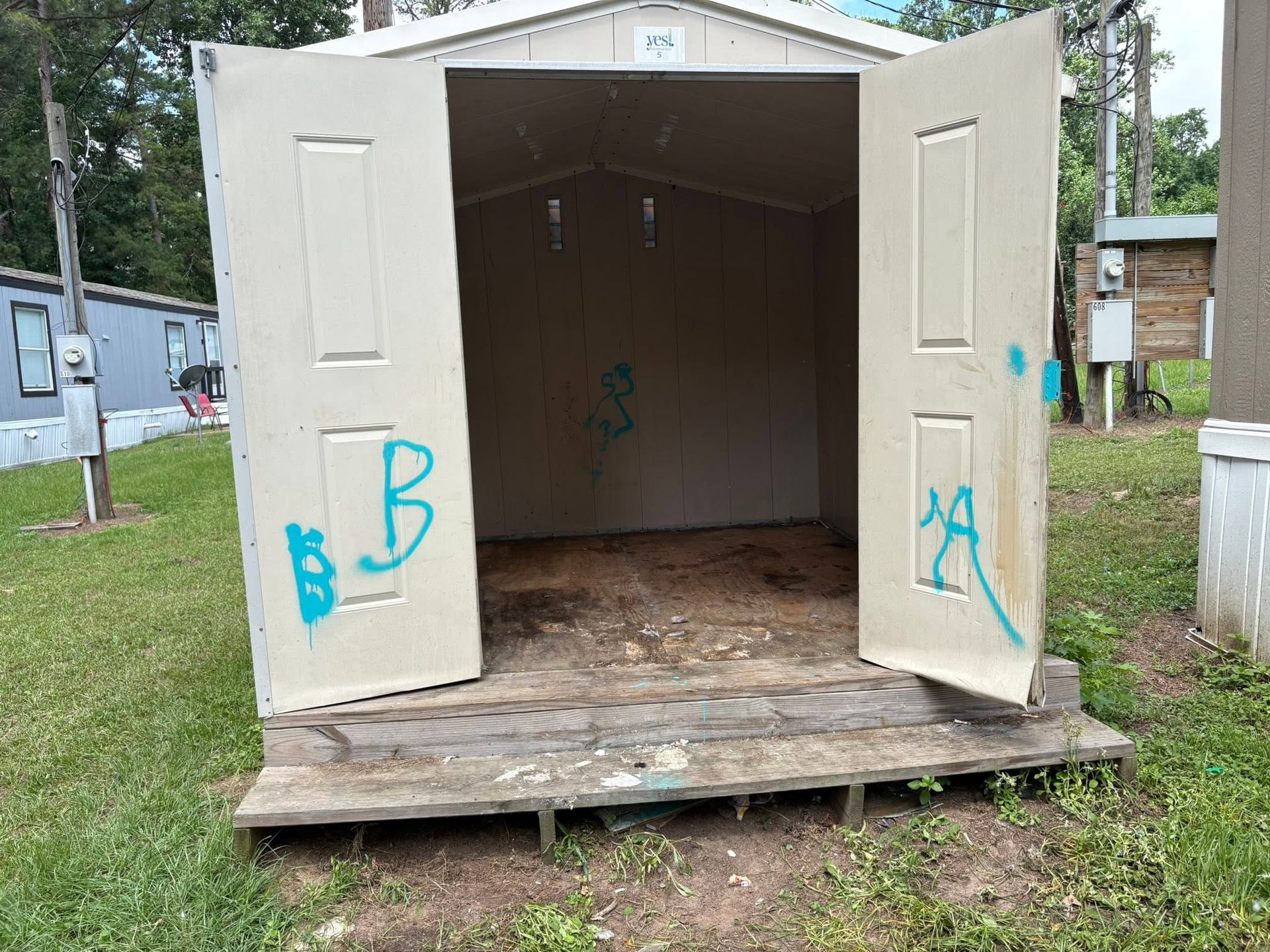 Open white shed with double doors standing on a grassy lot, with blue graffiti on the doors