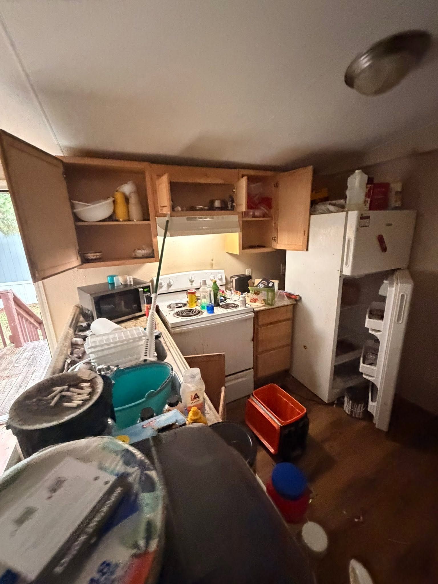 Cluttered kitchen with open cabinets, dishes piled in the sink, and trash bins on the floor.
