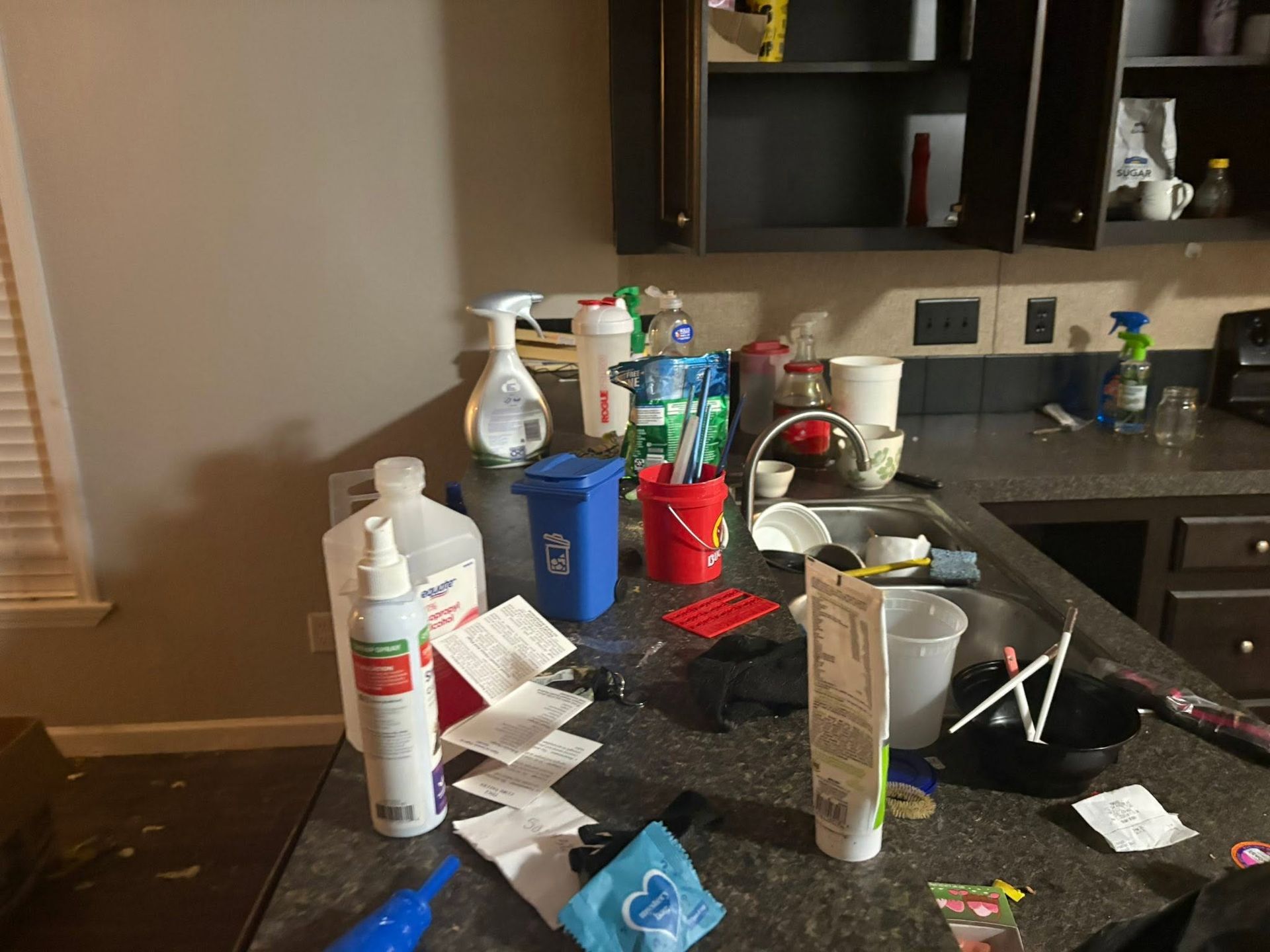 Cluttered kitchen counter with cleaning supplies, papers, cups, and dishes near a sink.