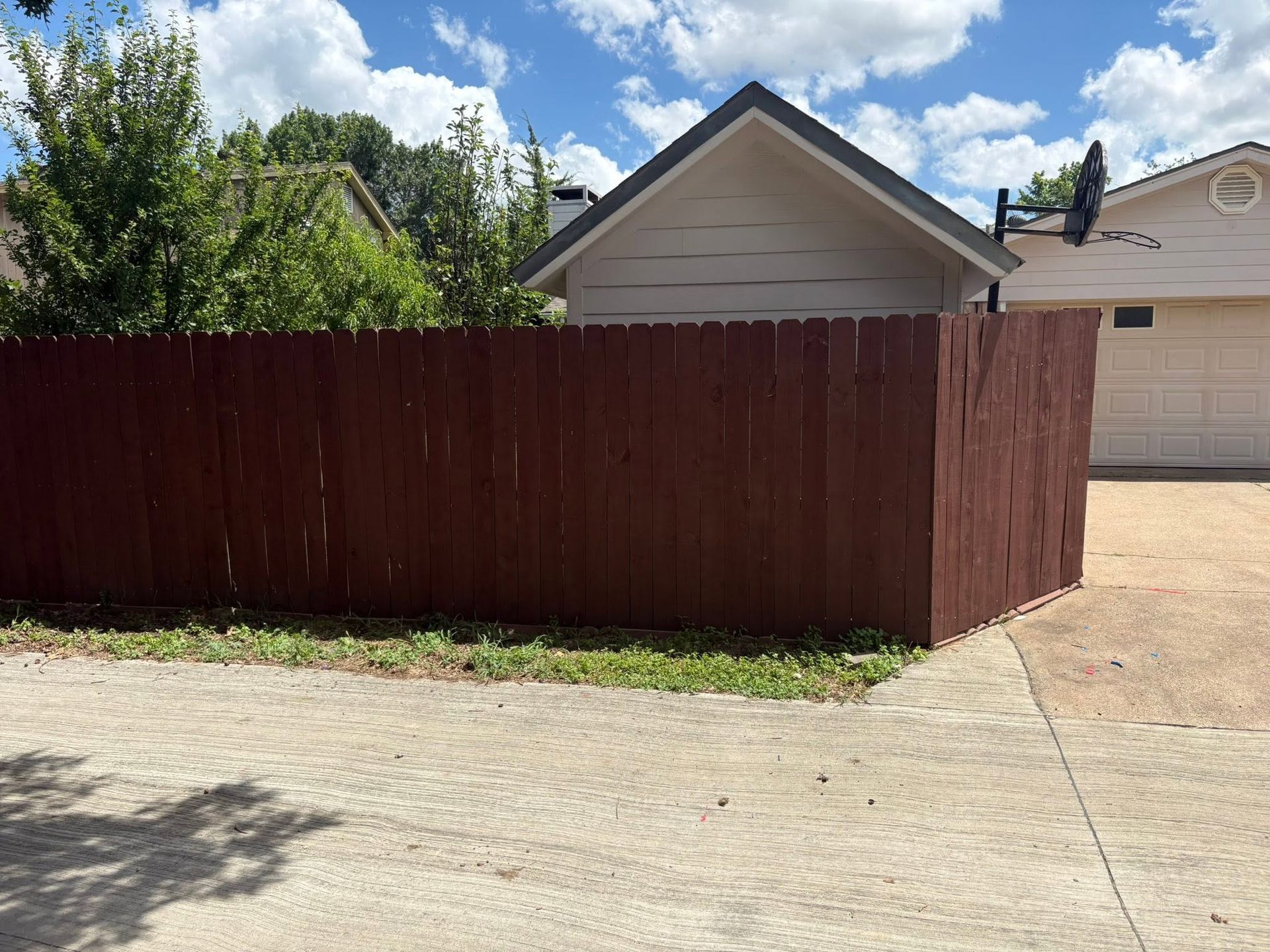 Red wooden fence beside a driveway, with a house and blue sky in the background.