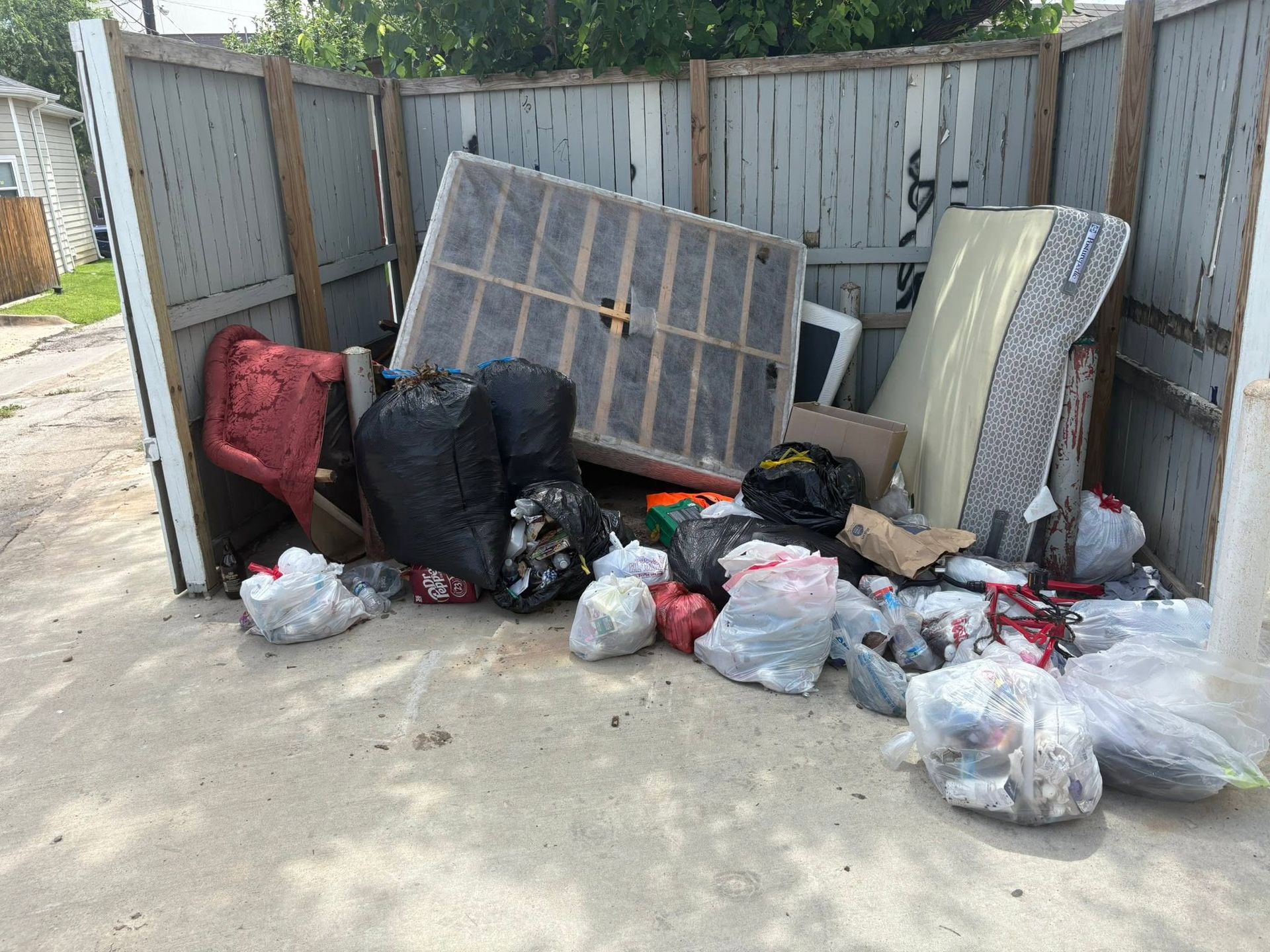 Overflowing dumpster area with trash bags, a mattress, and debris behind a metal fence.