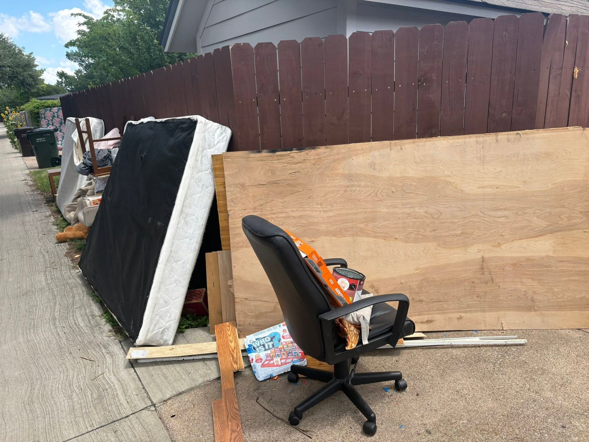 Black office chair and mattress leaning against a wooden fence in a driveway or alley
