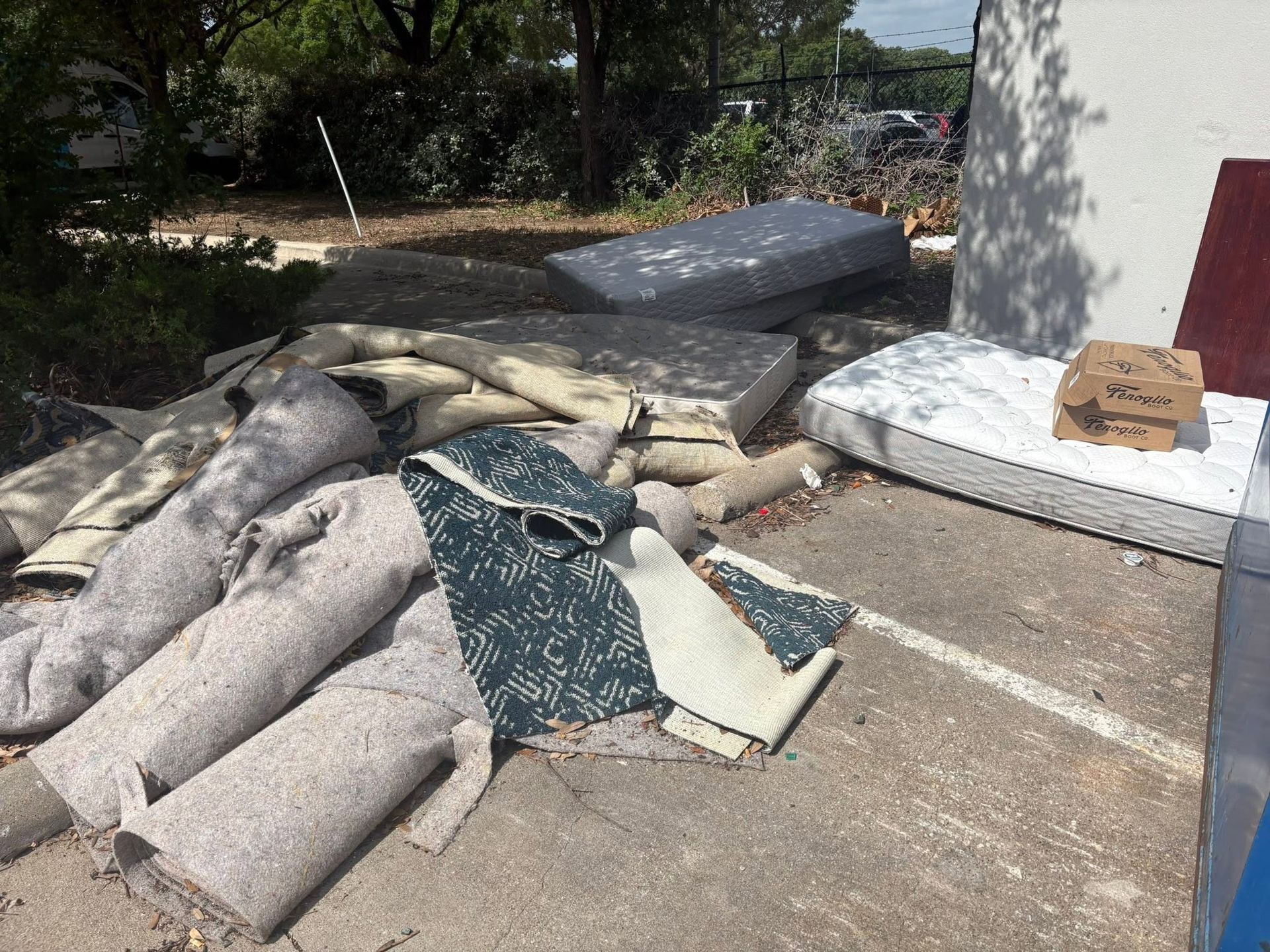 Pile of discarded mattresses and rolled carpet on a driveway beside a house and trees
